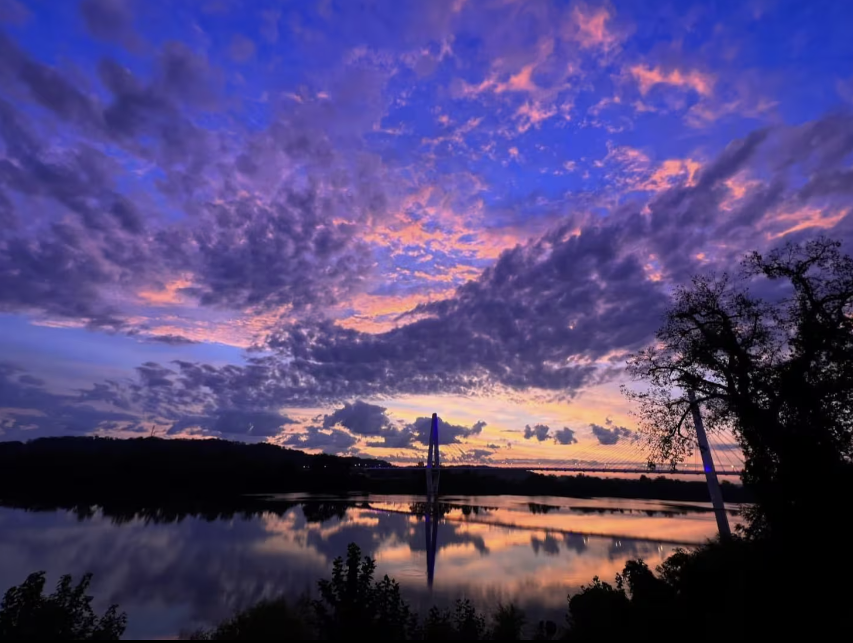 Colorful sunset over a river with a bridge and trees.