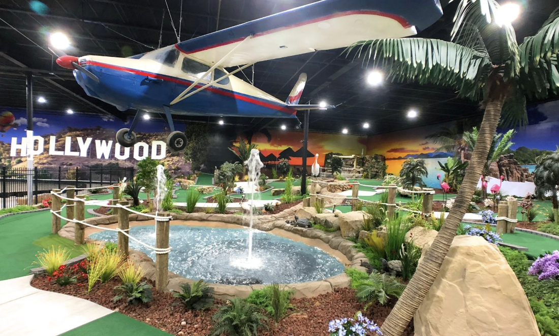 Indoor miniature golf course with a Hollywood theme, featuring a large airplane hanging from the ceiling, a water fountain, trees, bushes, and decorative plants, with a painted Hollywood sign and sunset in the background.