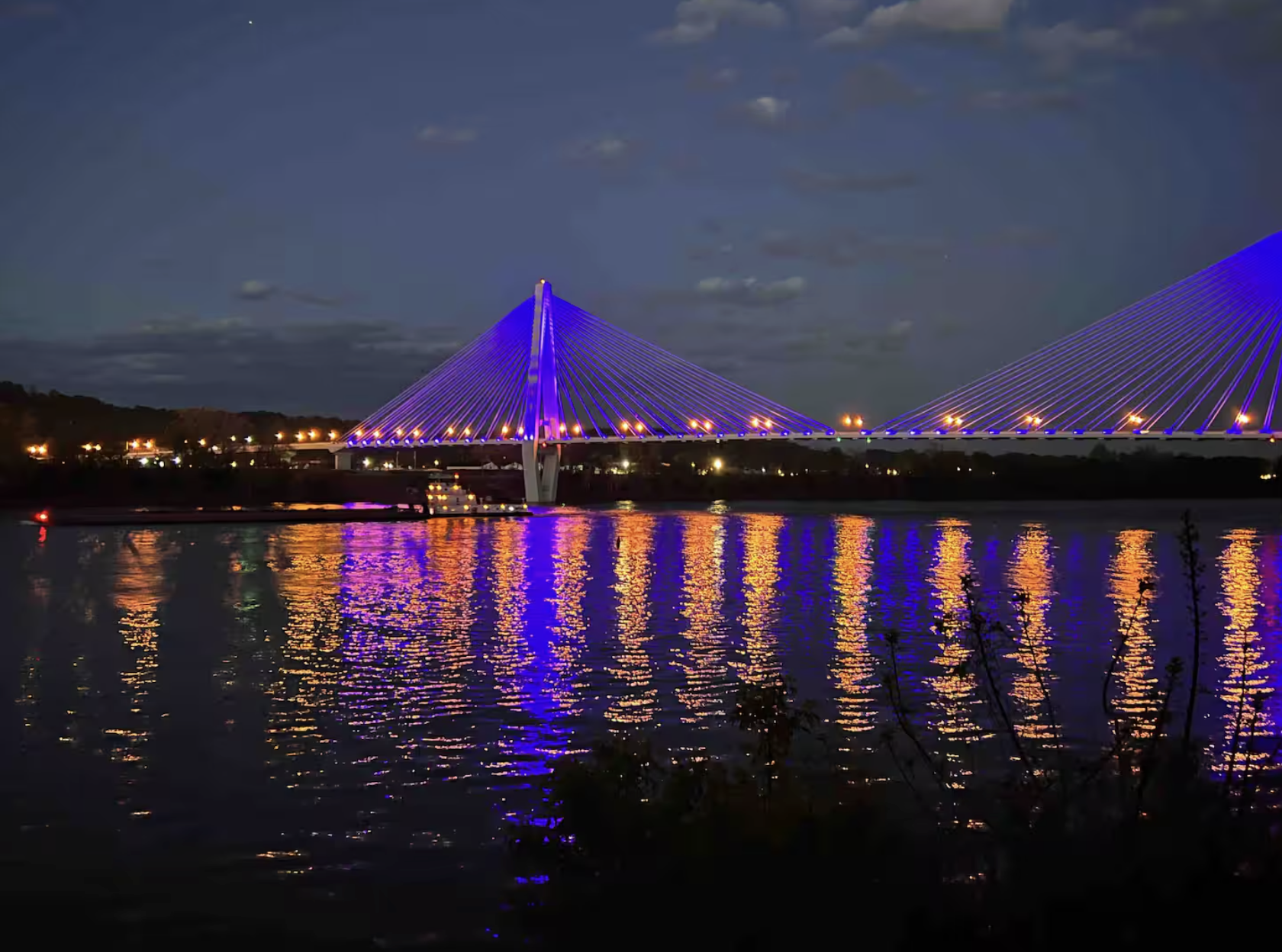 A brightly lit modern suspension bridge with blue and purple LED lights reflecting on a dark river, with a cloudy sky overhead. Small buildings with lights are visible in the background.