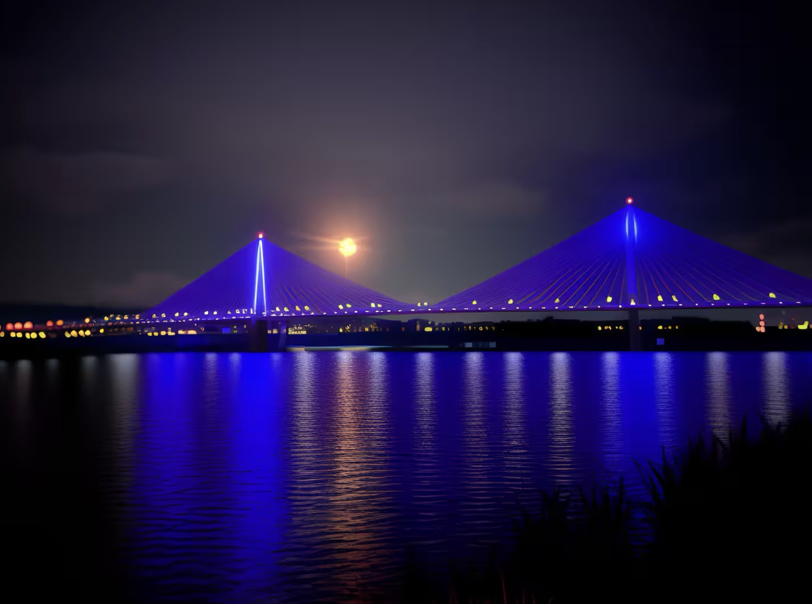 Night view of a bridge illuminated in blue lights reflected on a body of water, with city lights in the background and a full moon in the sky.
