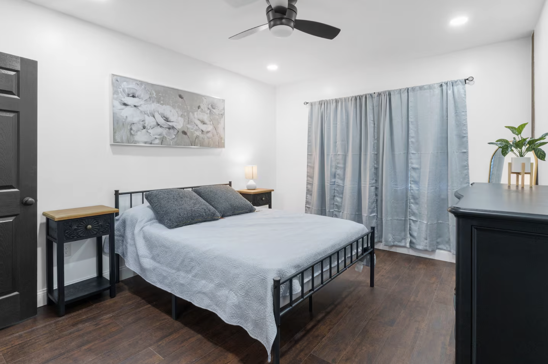 Bright bedroom with a black metal bed frame, white bedspread, and two dark gray pillows. A nightstand with a lamp is beside the bed. Light blue curtains cover a window, and a dresser with a plant is nearby. A ceiling fan with black blades is on the c