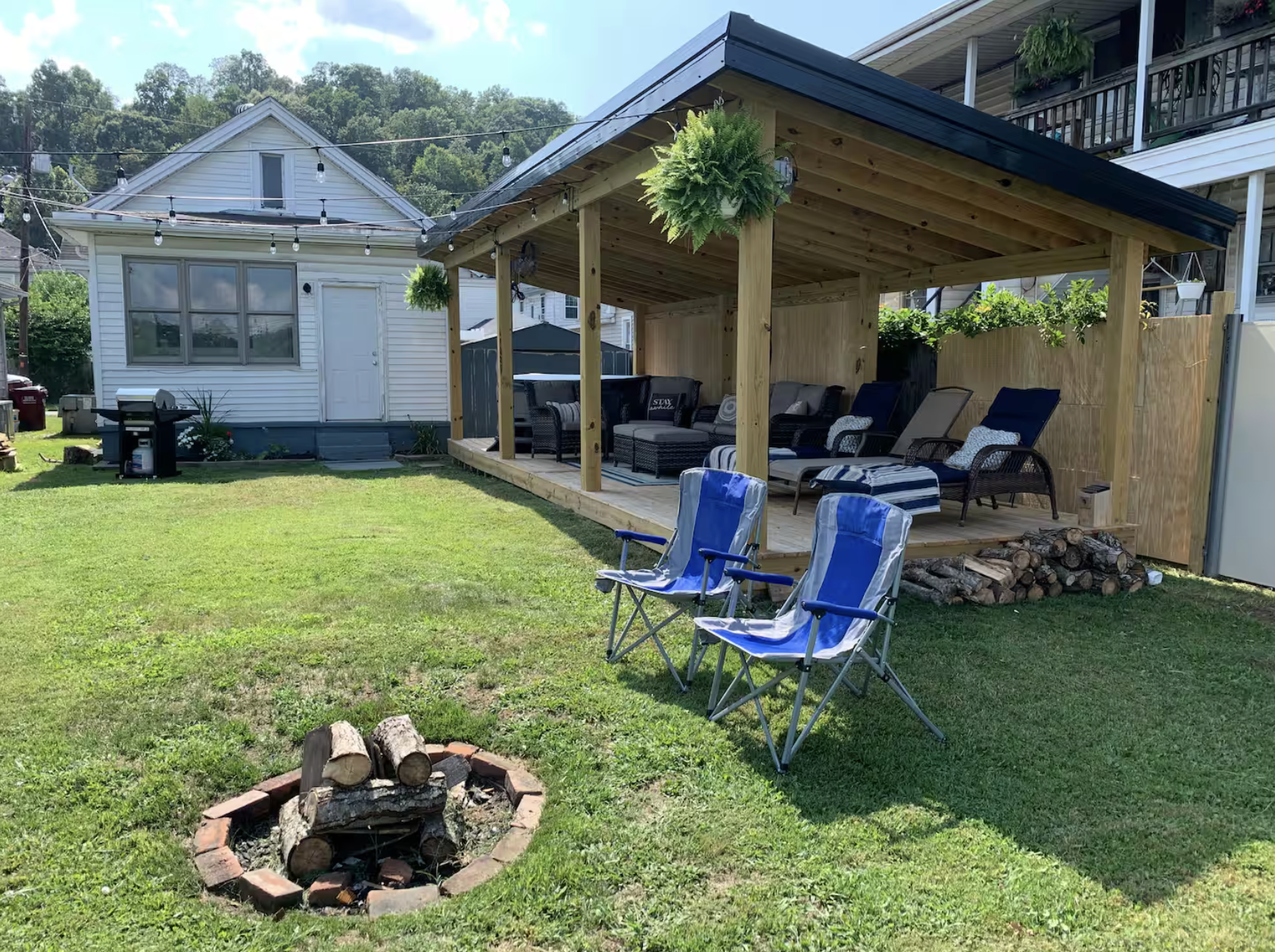 Backyard with a grassy area, a fire pit made of bricks, two blue folding chairs, a covered patio with outdoor furniture, hanging plants, a woodpile, a white house with string lights, and a fence with greenery.