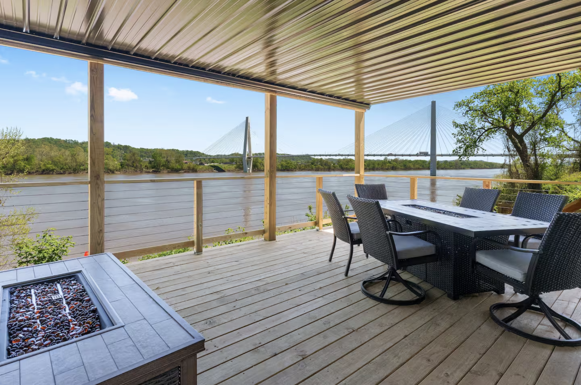 Covered wooden deck overlooking a river with a modern cable-stayed bridge in the background. The deck has a dining table with six chairs and a fire pit inset in the tabletop.
