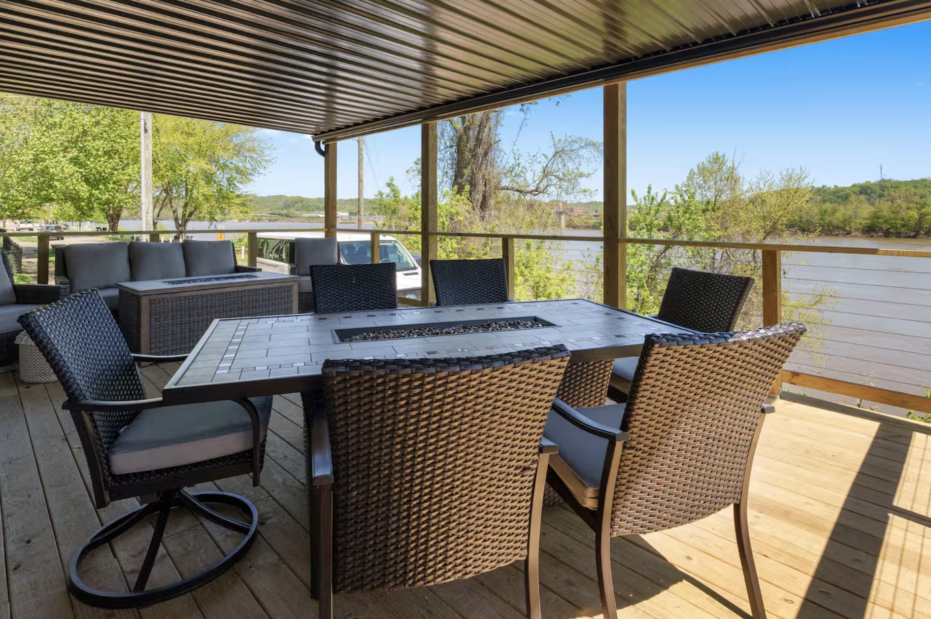 Outdoor patio with a dining table and six chairs overlooking a river and green trees under a partly cloudy sky.