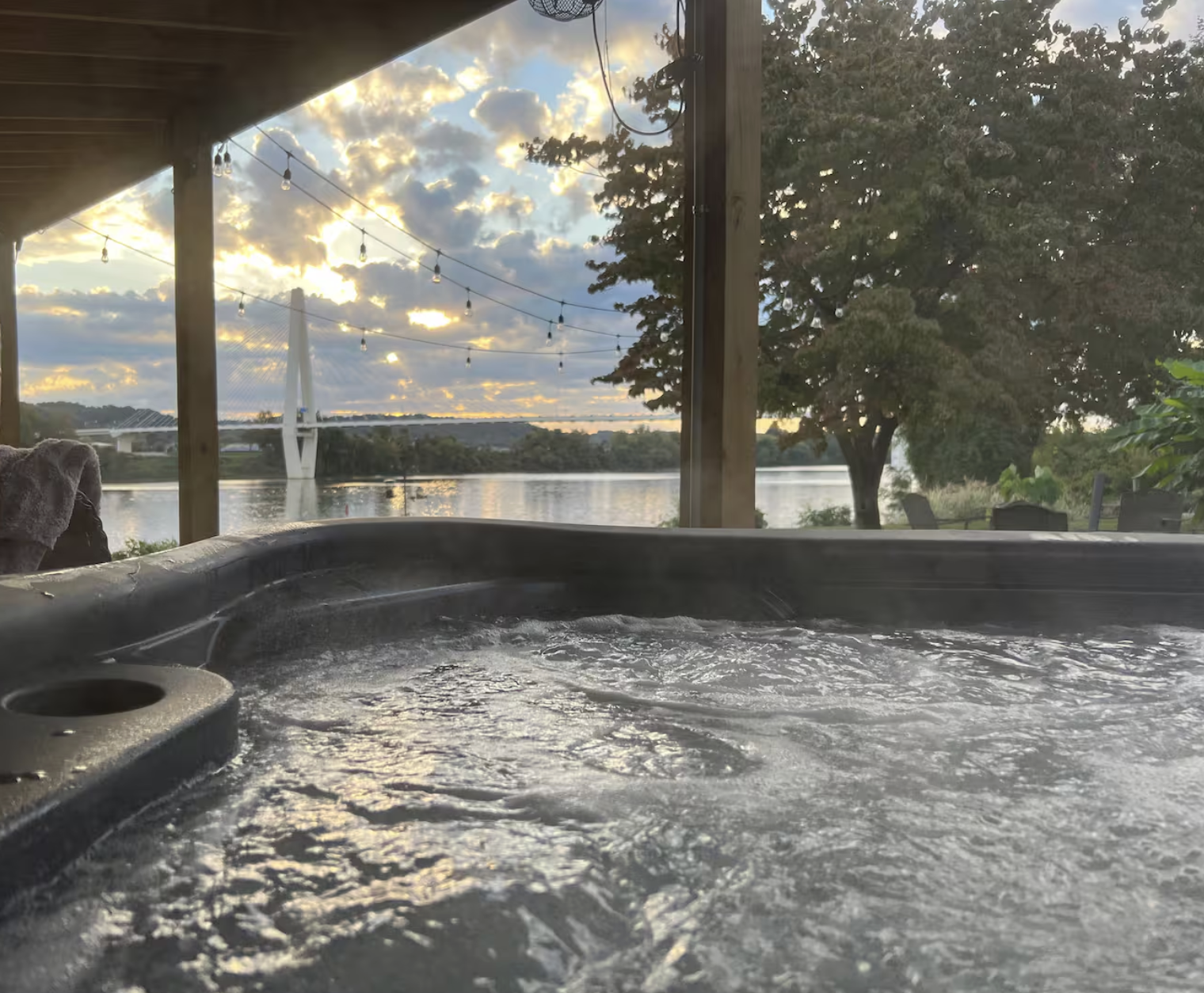 Hot tub on an outdoor deck overlooking a river with a bridge in the distance, trees, and a partly cloudy sky during sunset.