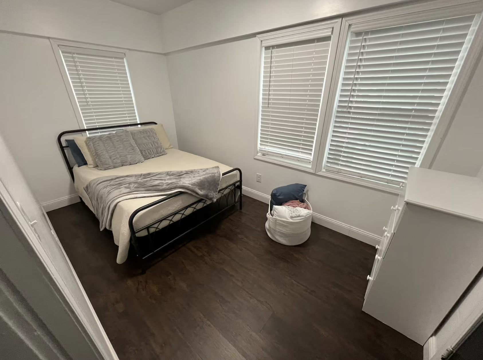 A minimalist bedroom with dark wooden floor, white walls, a black metal bed with grey and beige bedding and pillows, and three windows with white blinds.