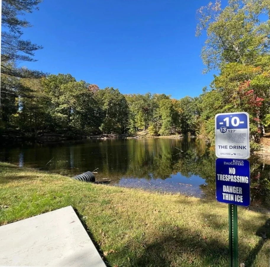 A lakeside scene with a clear blue sky, trees with green and some autumn-colored leaves, and a small lake with calm water. There is a sign on a post that indicates a water level of -10 feet, a warning against trespassing, and thin ice danger.
