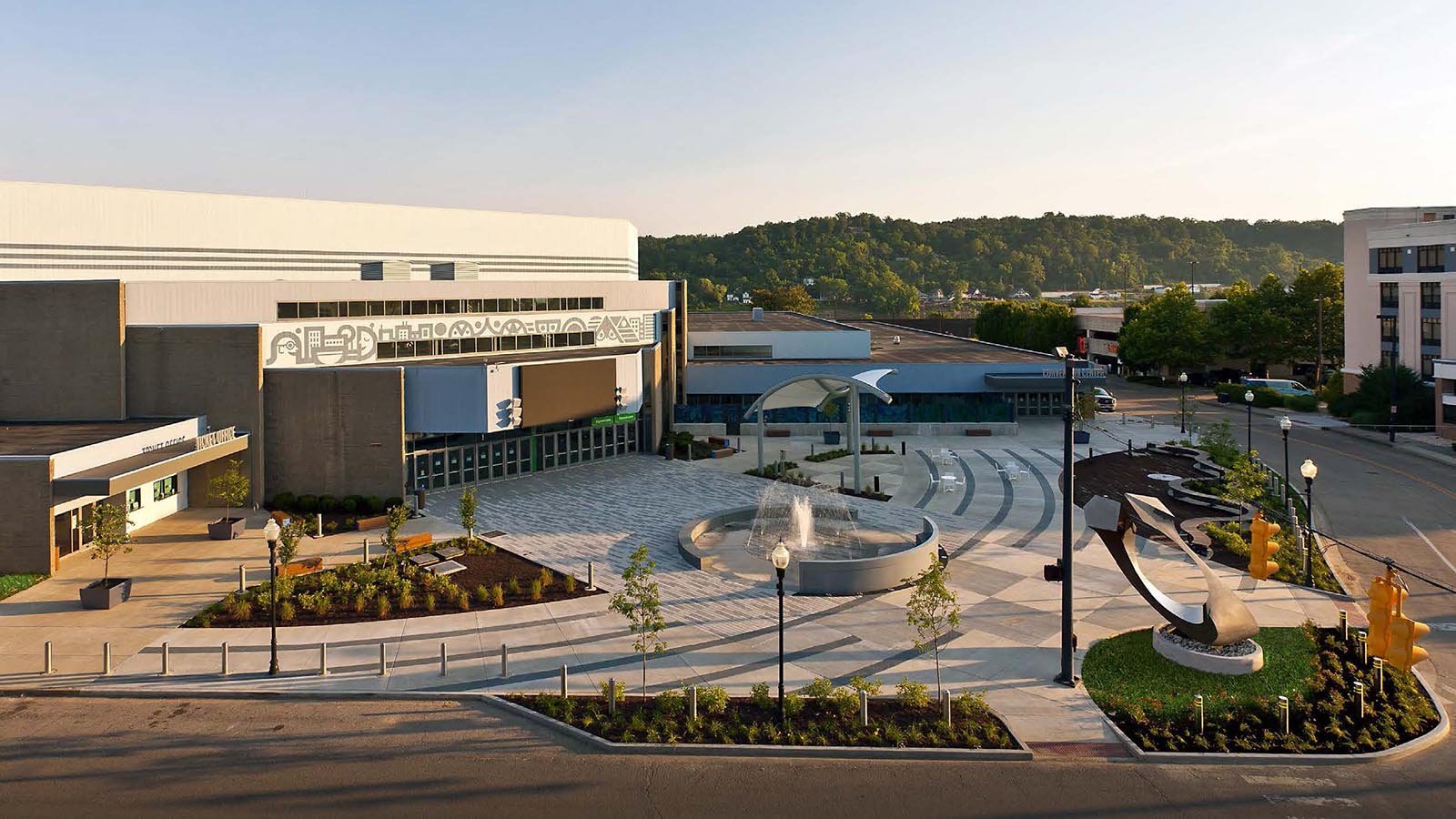 An aerial view of a modern downtown area with a large public square featuring a fountain, sculptures, trees, and lampposts surrounded by commercial and office buildings with a cityscape and green hills in the background.