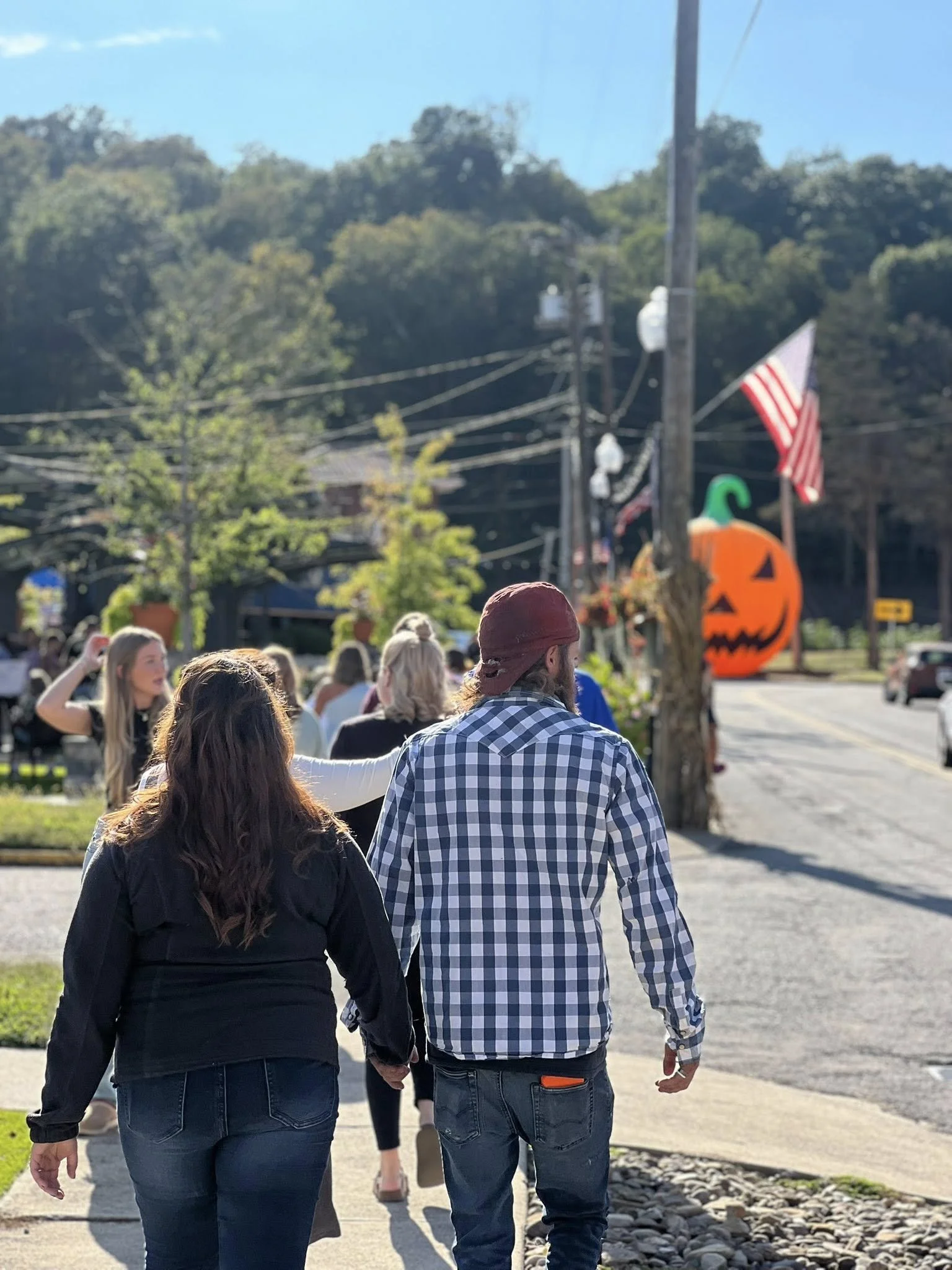 A couple walking hand in hand outdoors with a crowd, Halloween decorations including a large pumpkin with a green stem and an American flag in the background.