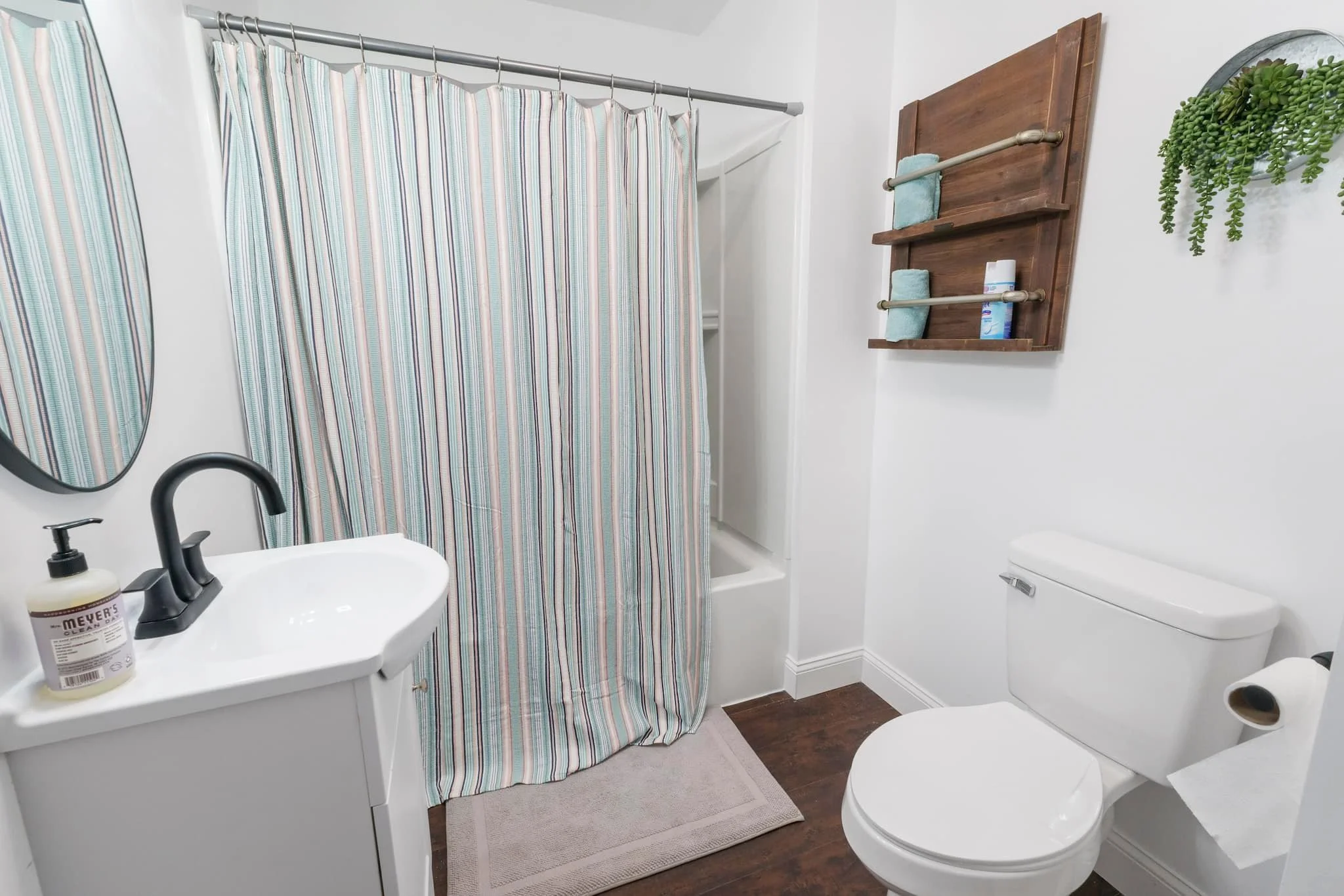A small bathroom with a white toilet, a white sink with a black faucet, a striped shower curtain, a wooden wall shelf with towels and toiletries, a mirror, and a small plant on the wall.