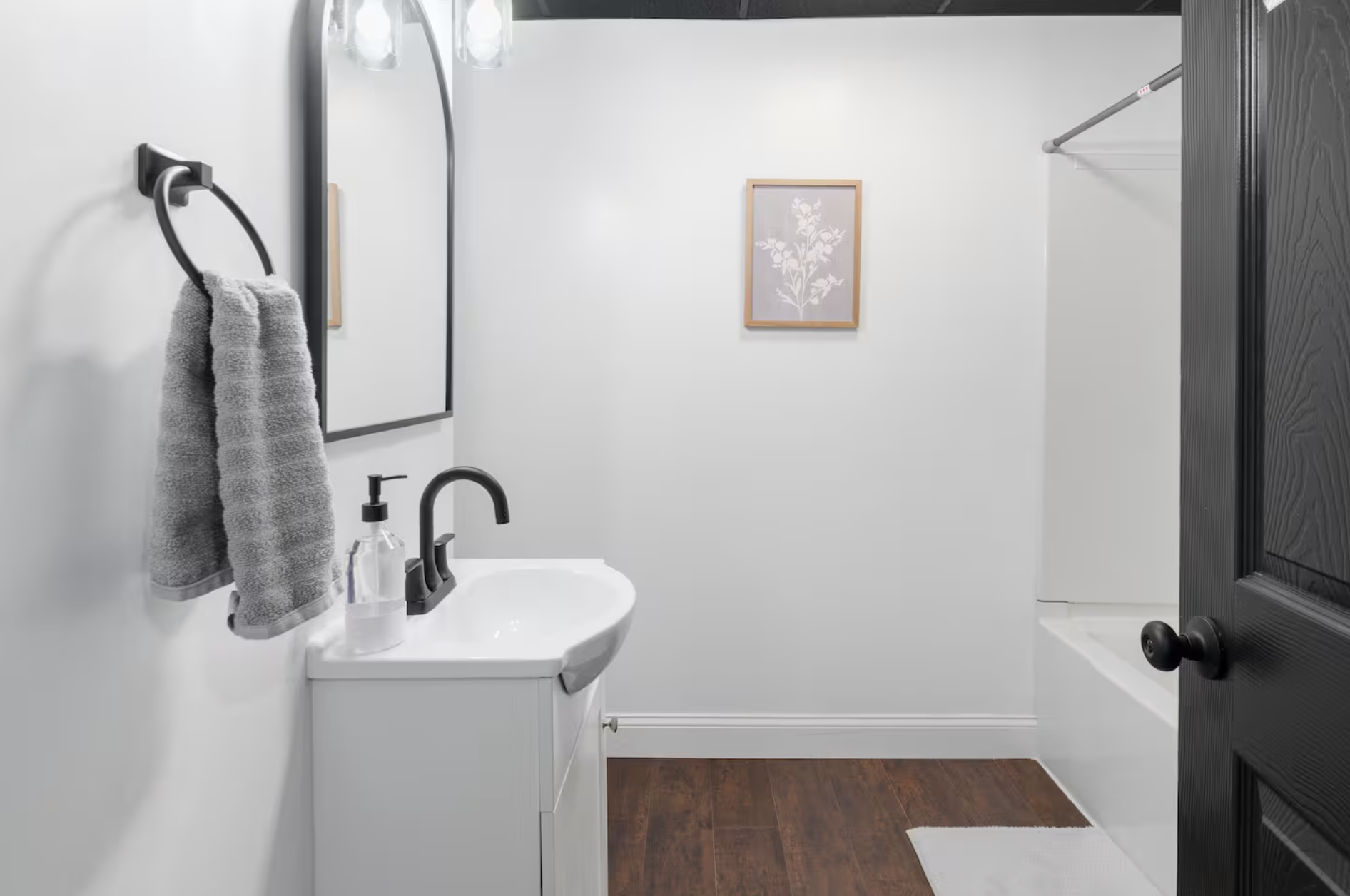 A bathroom with white walls, a black-framed mirror, a small white vanity with a black faucet, a gray towel hanging on a black towel ring, a framed botanical print on the wall, a partial view of a shower with a black rod, and dark wood flooring.