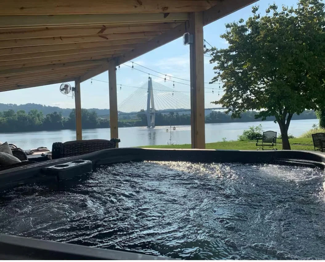 Hot tub on a covered patio overlooking a river with a bridge, trees, and outdoor chairs.