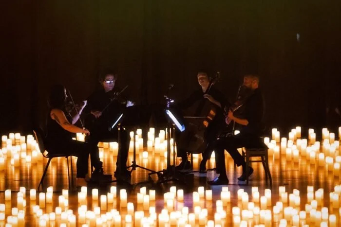 Four musicians playing string instruments in a dark room surrounded by numerous lit candles on the floor.