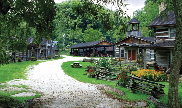 A rural scene with dirt paths, old wooden buildings, a covered porch, and a garden with flowers and fences, set against a backdrop of green trees and hills.