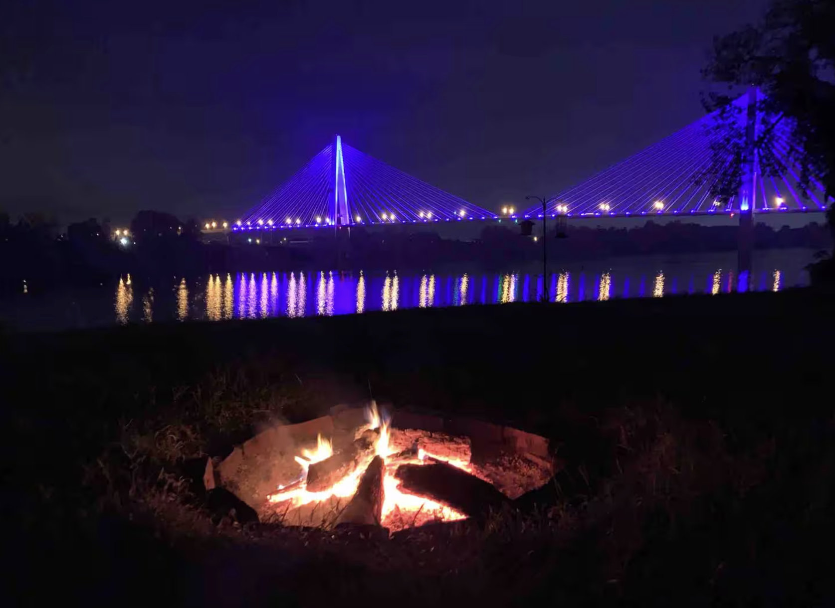 Night scene of a bridge illuminated with purple lights reflecting on the water, with a small campfire burning in the foreground.