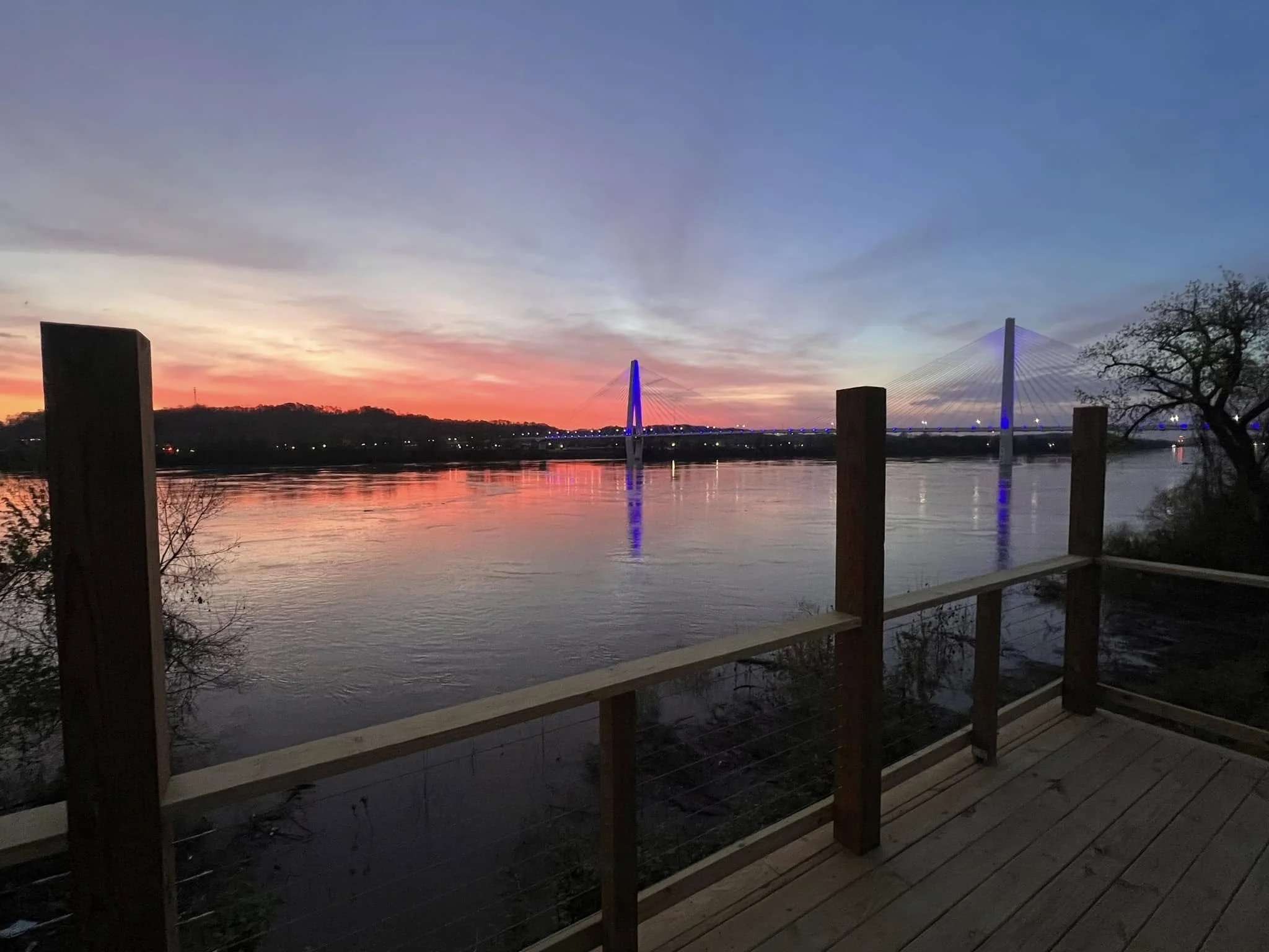 A riverside view during sunset with a bridge illuminated with blue lights. The sky has hues of orange, pink, and purple, and the bridge's lights reflect on the calm water.