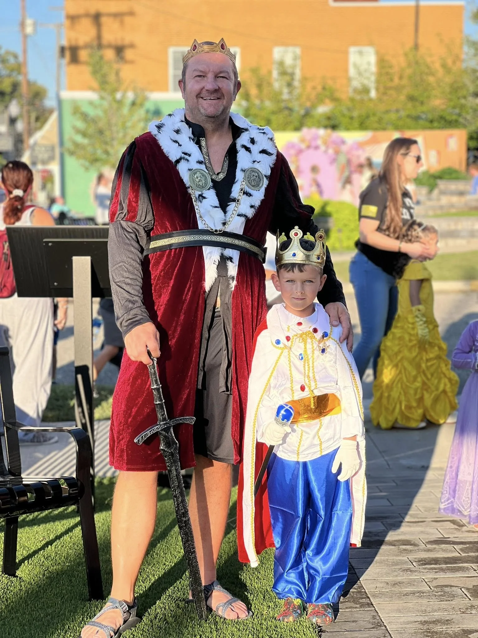 A man and a young boy dressed as a king and a prince, both wearing crowns, standing outdoors at a costume event. The man is dressed as a king with a crown, red robe with white fur trim, and holding a sword, while the boy is dressed as a prince with a crown, white cape, and blue pants.