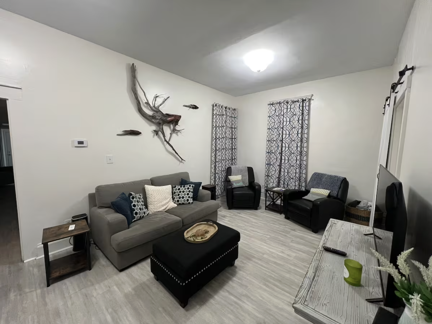 Living room with gray sofa, black and gray armchairs, patterned curtains, and wall decoration featuring a large driftwood piece and fish sculptures.