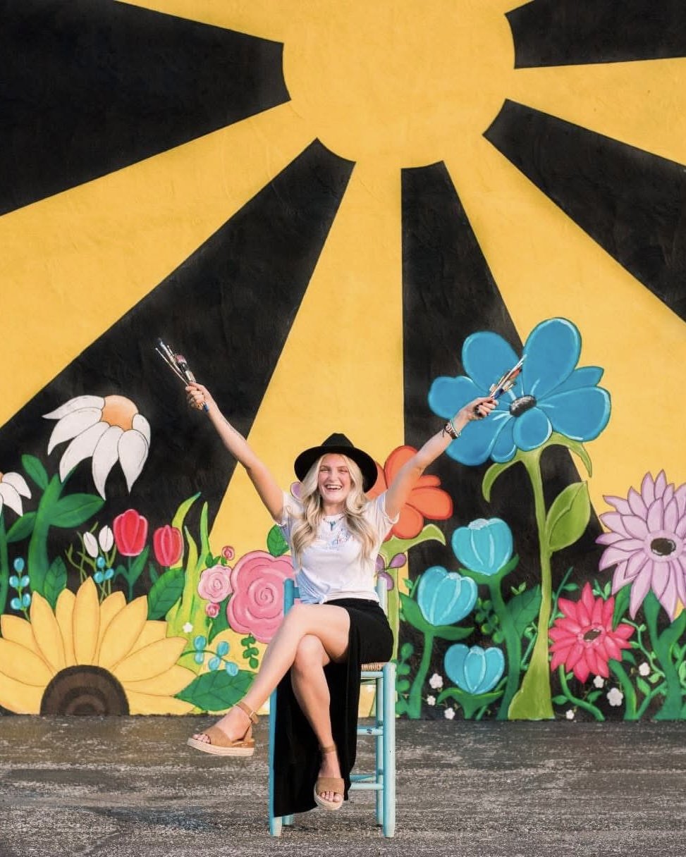 Woman sitting on a chair with her arms raised, holding sparklers, in front of a colorful mural of flowers and sun rays.
