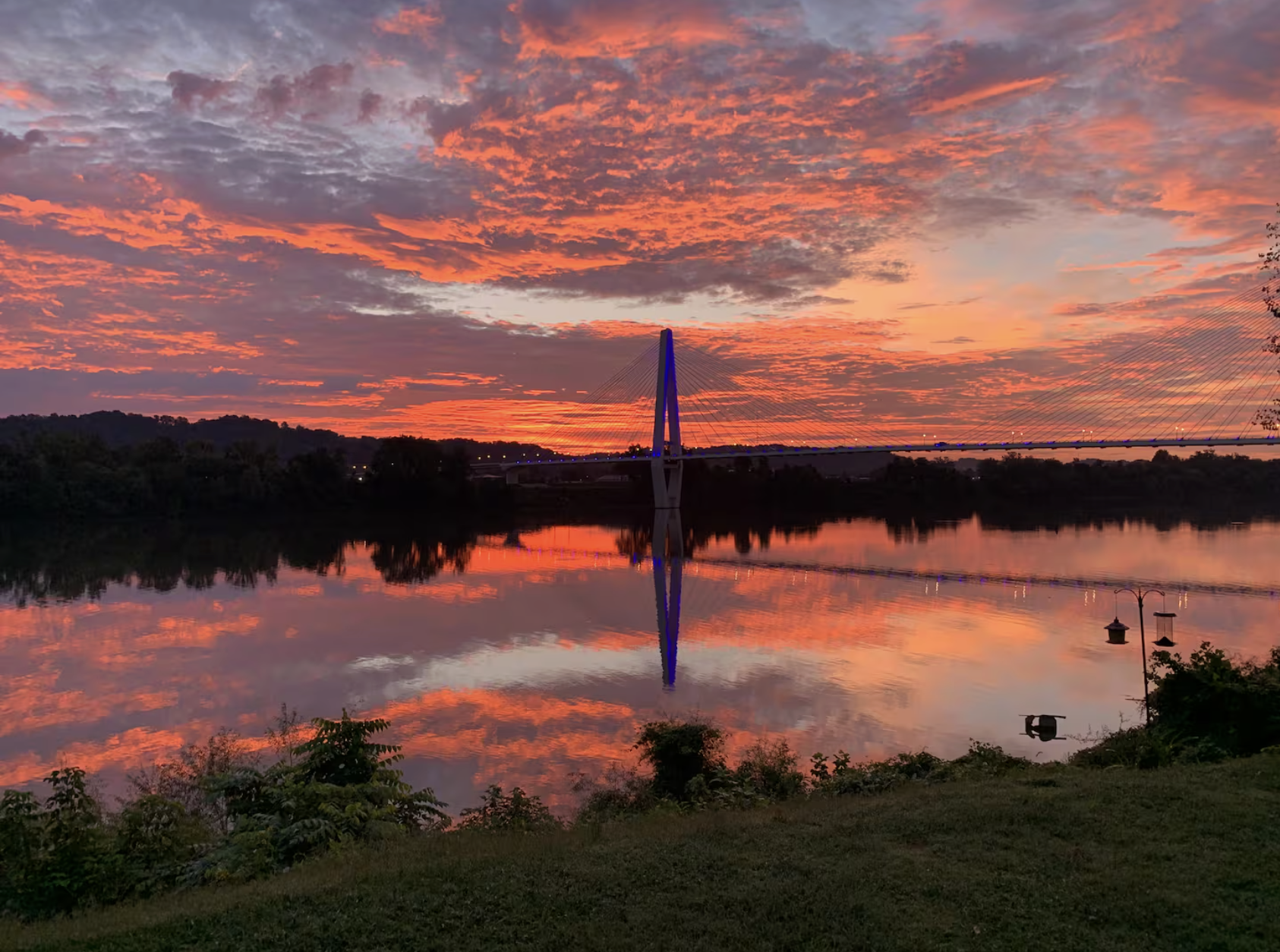 A colorful sunset sky with orange, pink, and purple clouds reflected on a calm river, with a bridge in the distance and some trees and bushes along the riverbank.