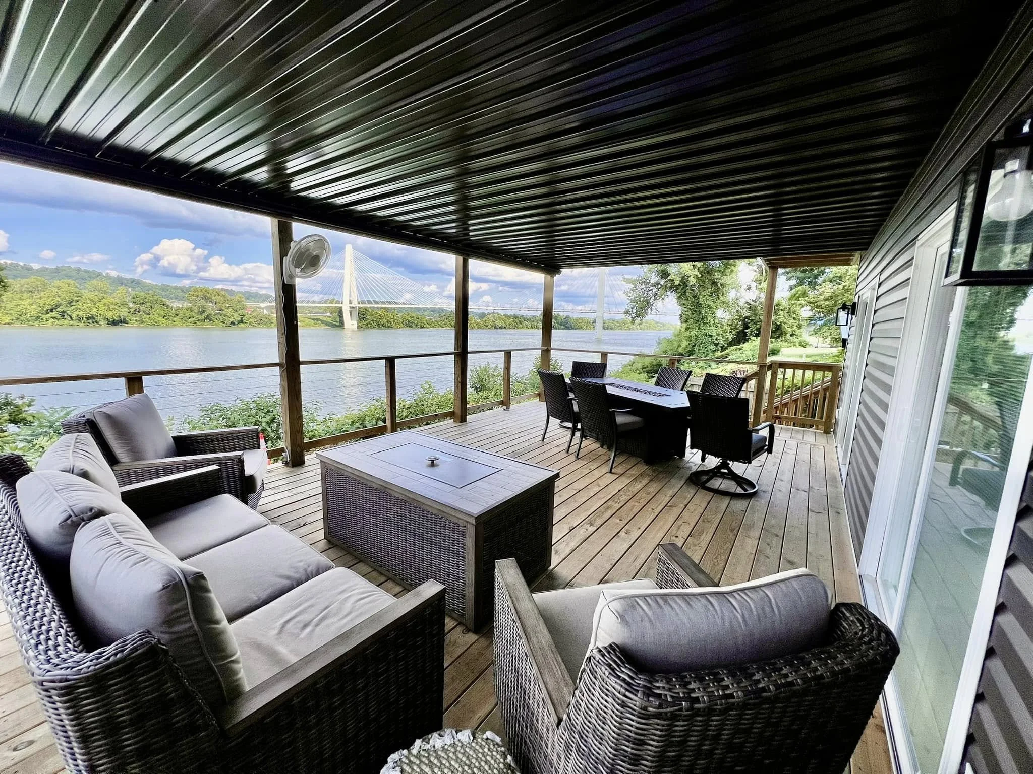 Covered outdoor deck with wicker furniture, a dining table, and a view of a river and a cable-stayed bridge in the distance.