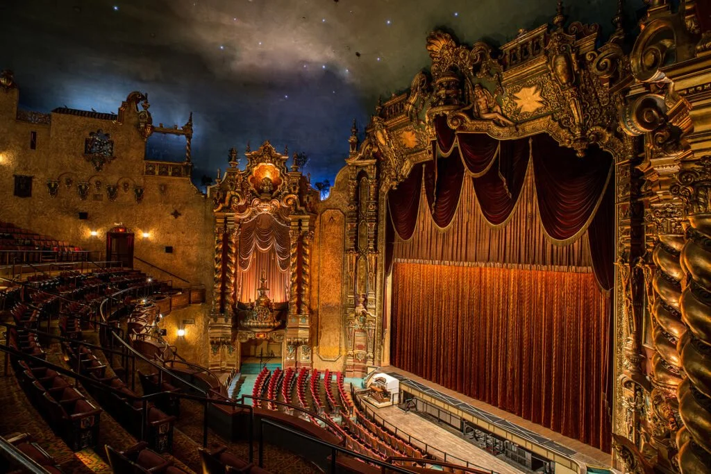 An ornate theater with a large stage covered by closed velvet curtains, elaborate gold and red decorations, and tiered seating.