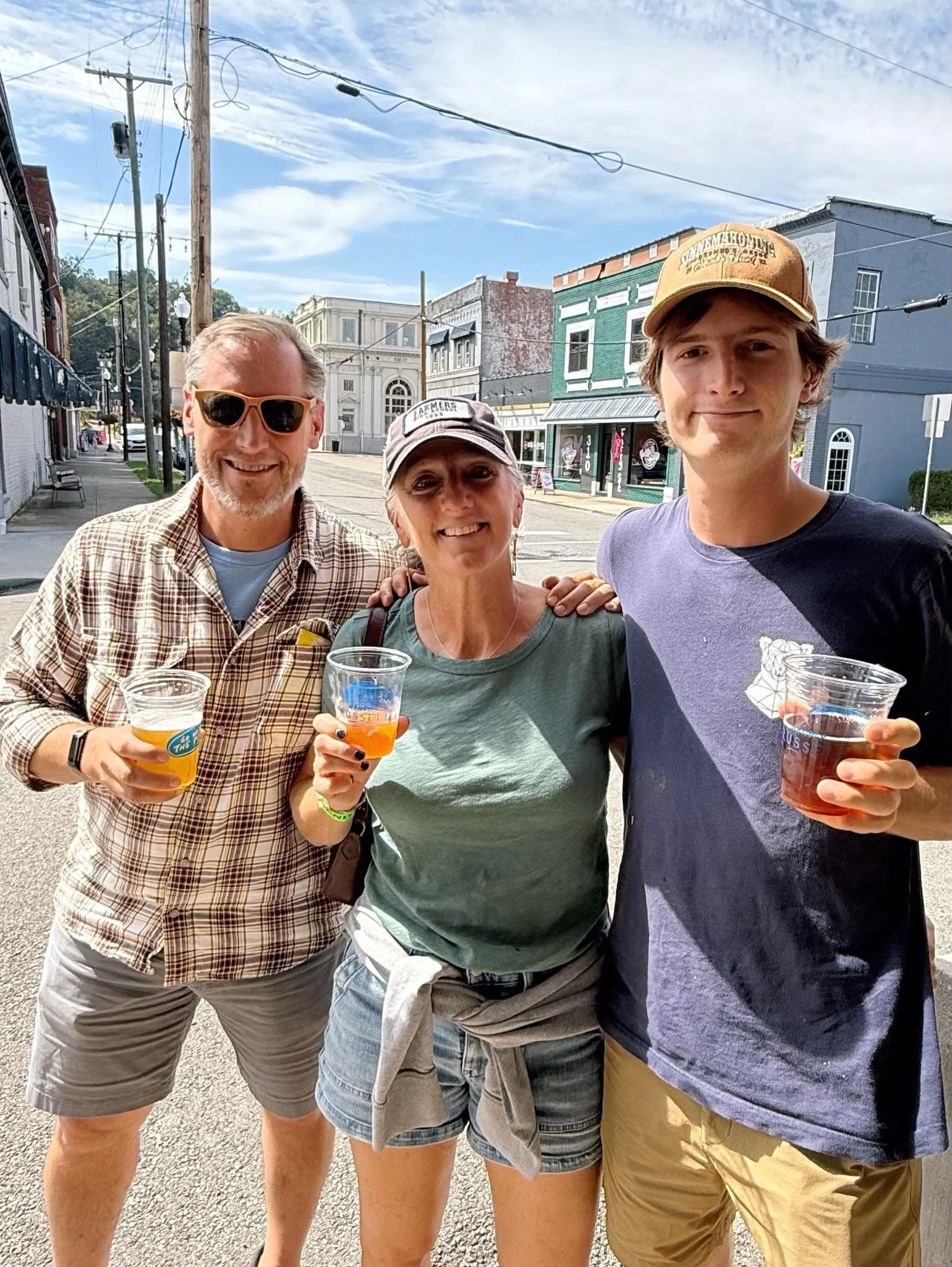 Three people standing on the street with drinks in plastic cups, smiling for the camera on a sunny day, with storefronts and utility poles in the background.
