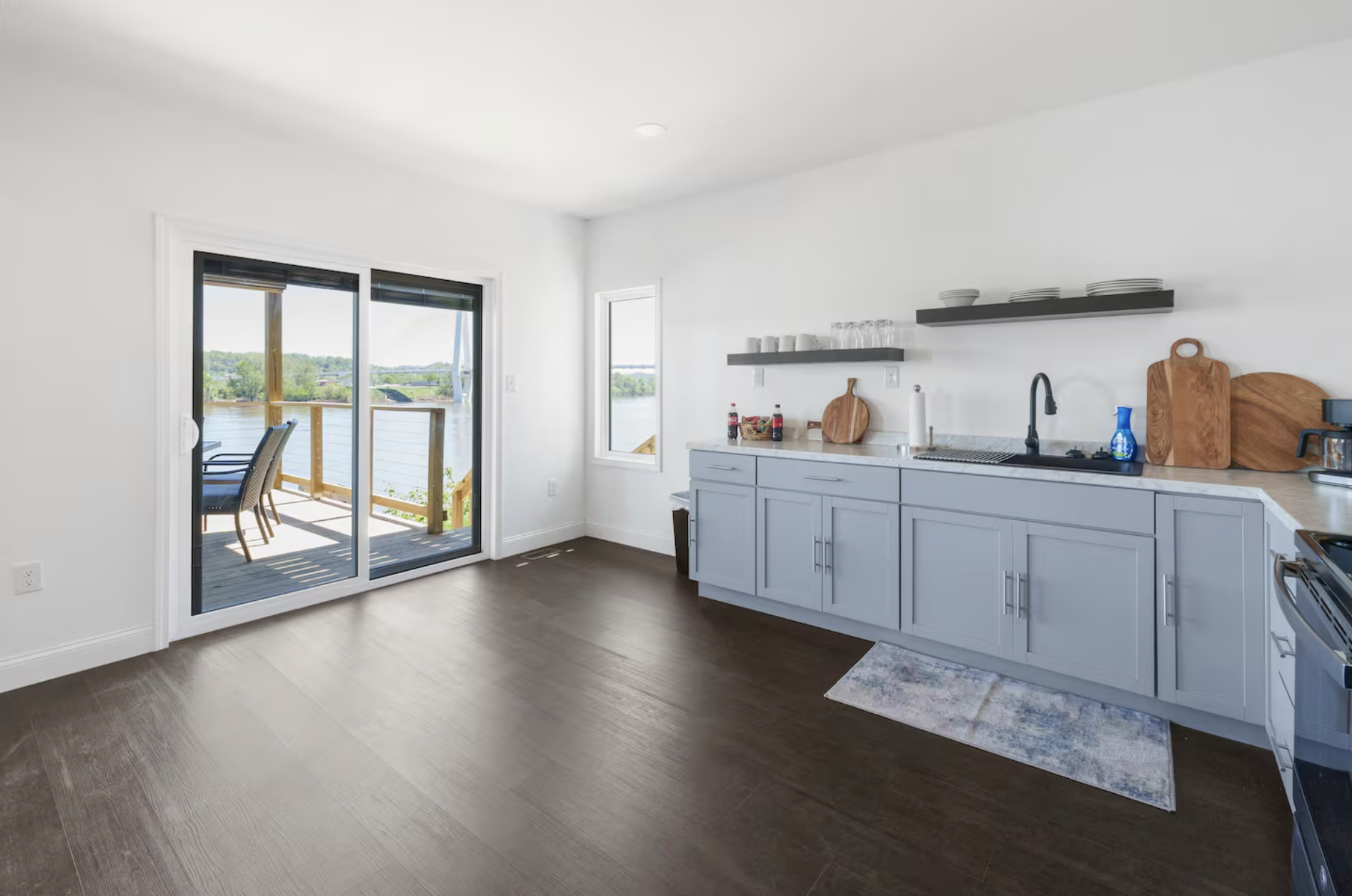 Empty kitchen with light blue cabinets, a dark wooden floor, a sliding glass door leading to a balcony with outdoor seating, and a window overlooking a body of water.