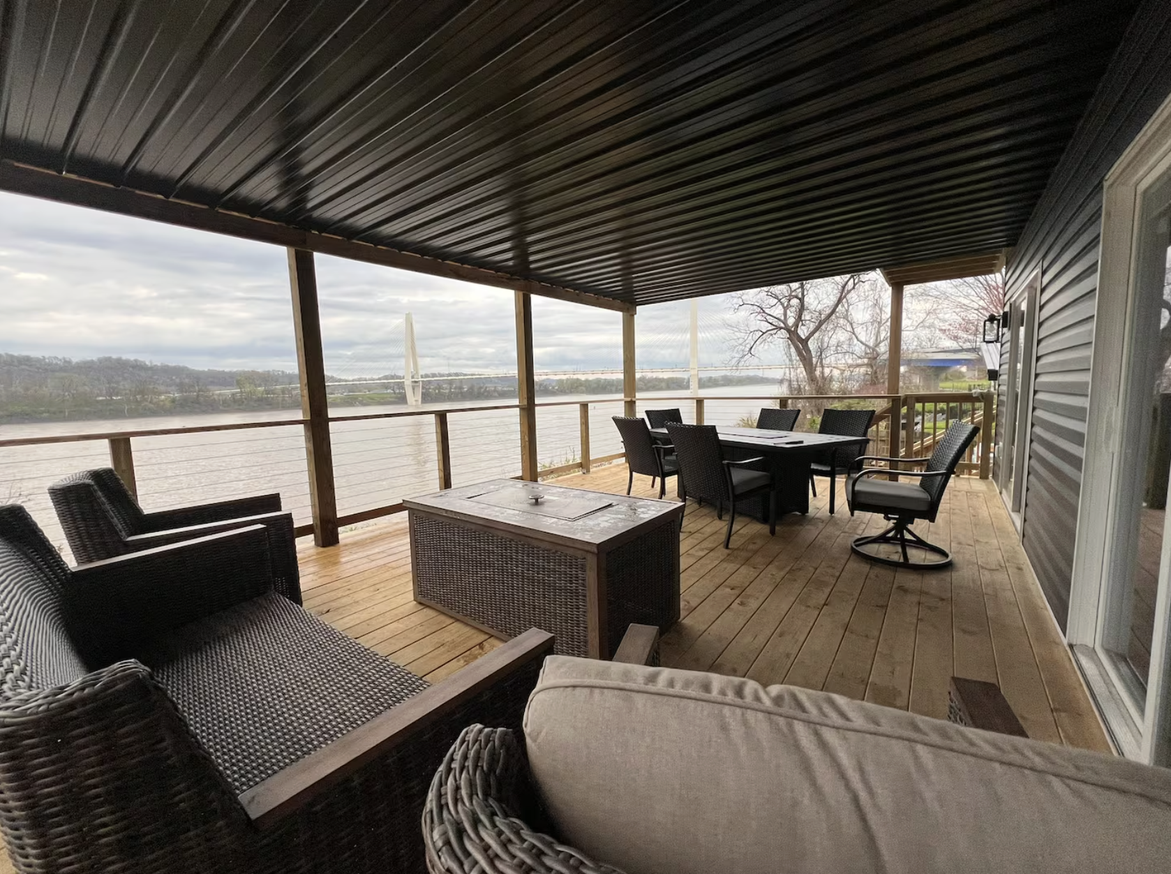 Covered wooden deck overlooking a river with outdoor furniture, including wicker chairs, a dining table with chairs, and a sun lounge. A bridge is visible in the distance over the water, and a tree without leaves is on the right.
