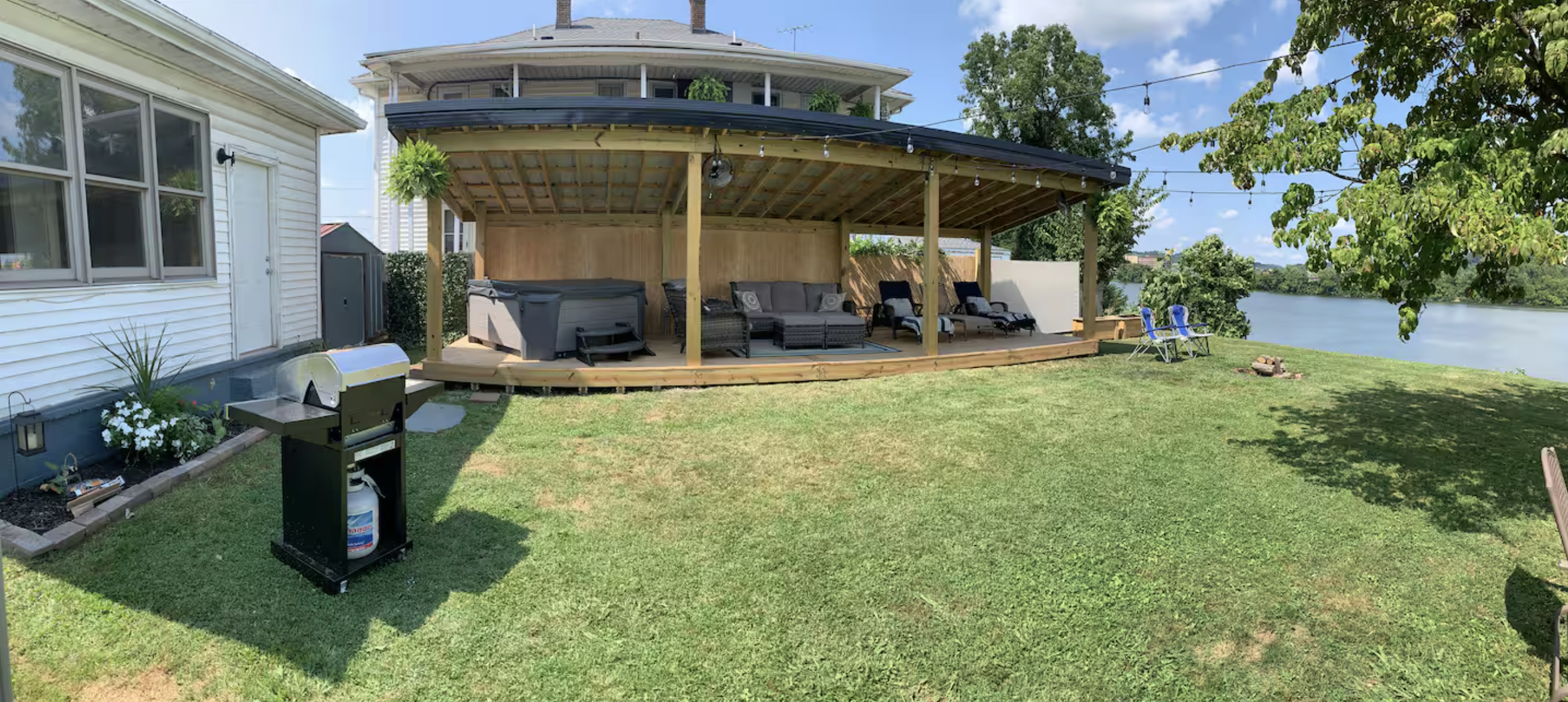 Backyard with a covered deck overlooking a river, outdoor furniture, chairs, a grill, and lawn with trees and blue sky.