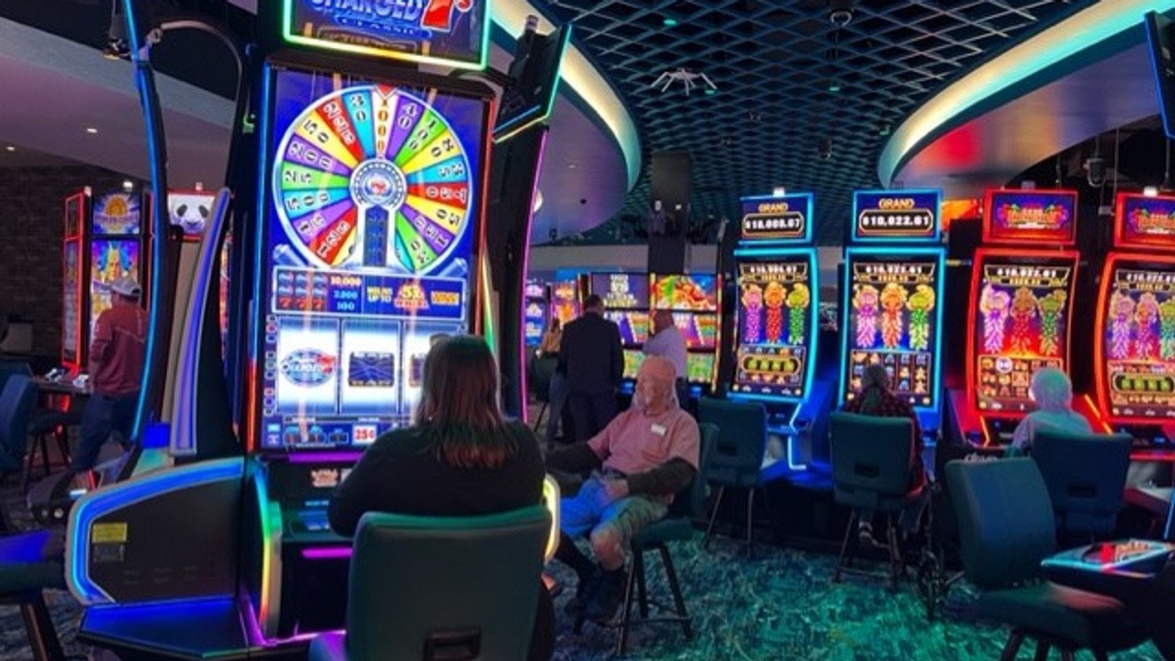 People playing slot machines in a brightly lit casino with colorful neon signs and a large spinning wheel game.