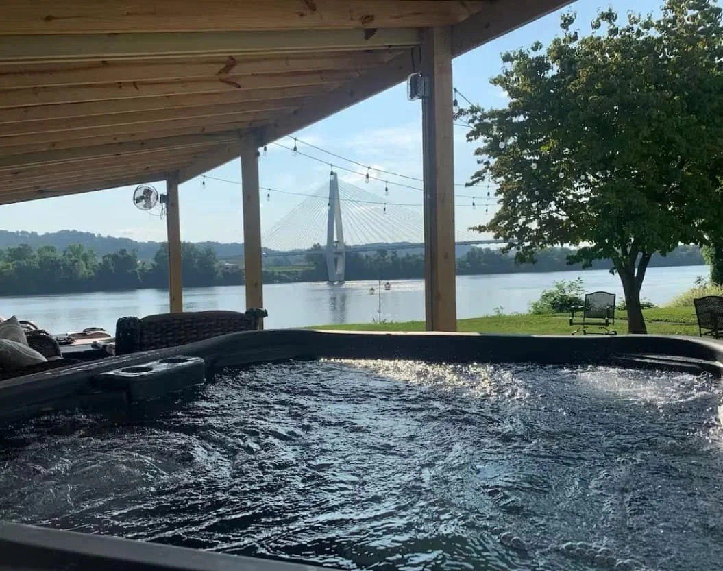 Hot tub on a covered patio overlooking a river with a bridge and trees in the background, with string lights hanging from the wooden ceiling.