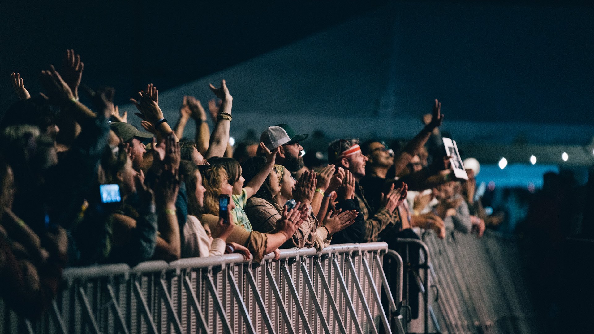 Audience at concert or event, standing behind a barricade, cheering and clapping with some holding phone, in a dark indoor setting.