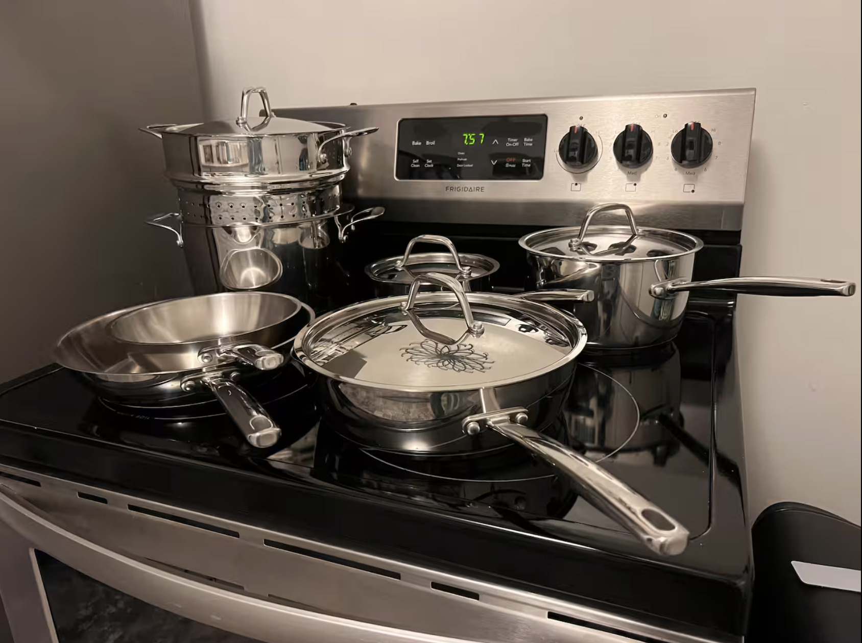 An electric stovetop with multiple stainless steel pots, pans, and strainers stacked and placed on burners, with a digital clock reading 7:57.