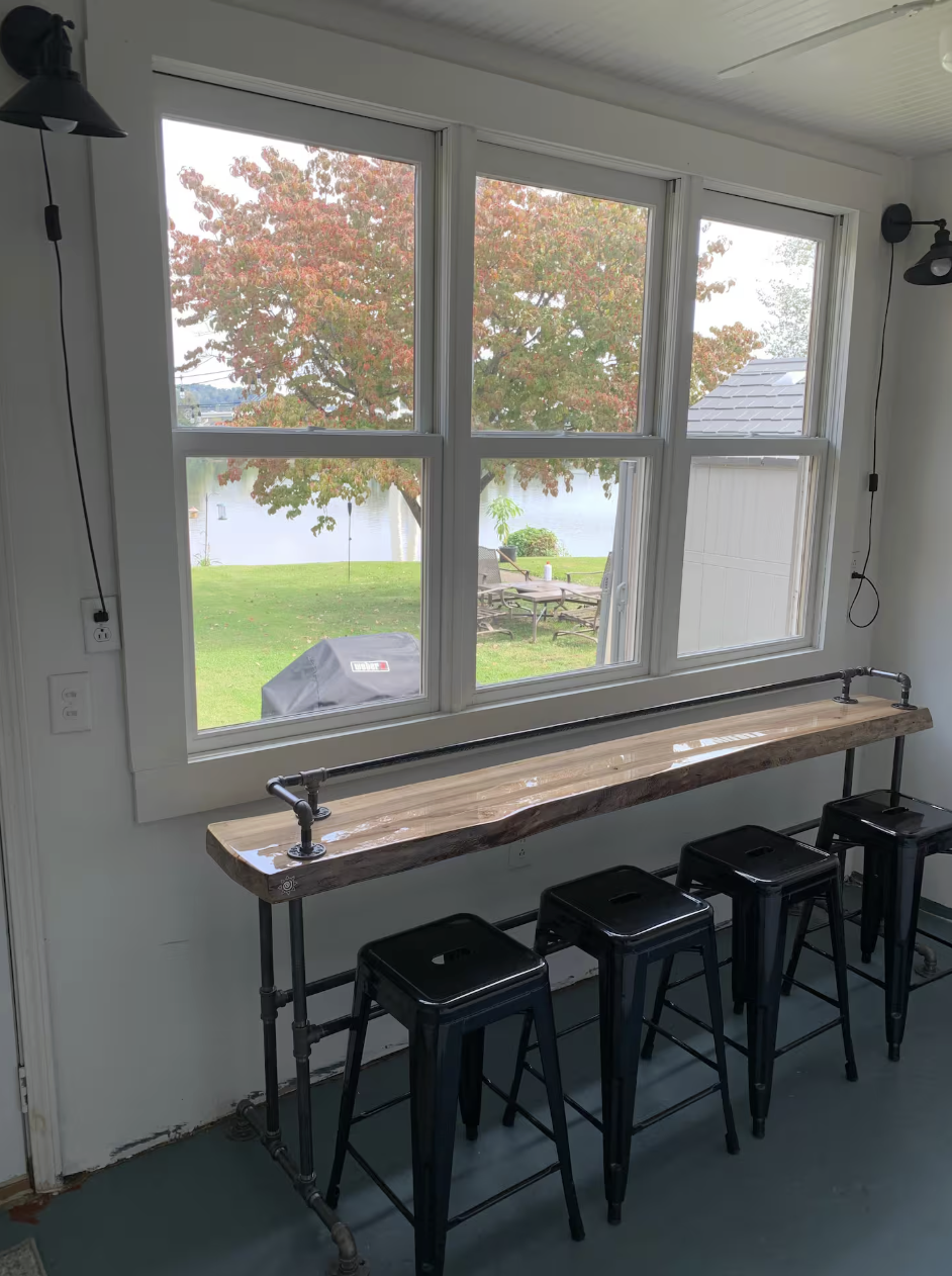 Interior view of a room with a large window overlooking a backyard with trees and outdoor furniture. There is a wooden bar-style table with industrial pipe legs and four black metal stools.