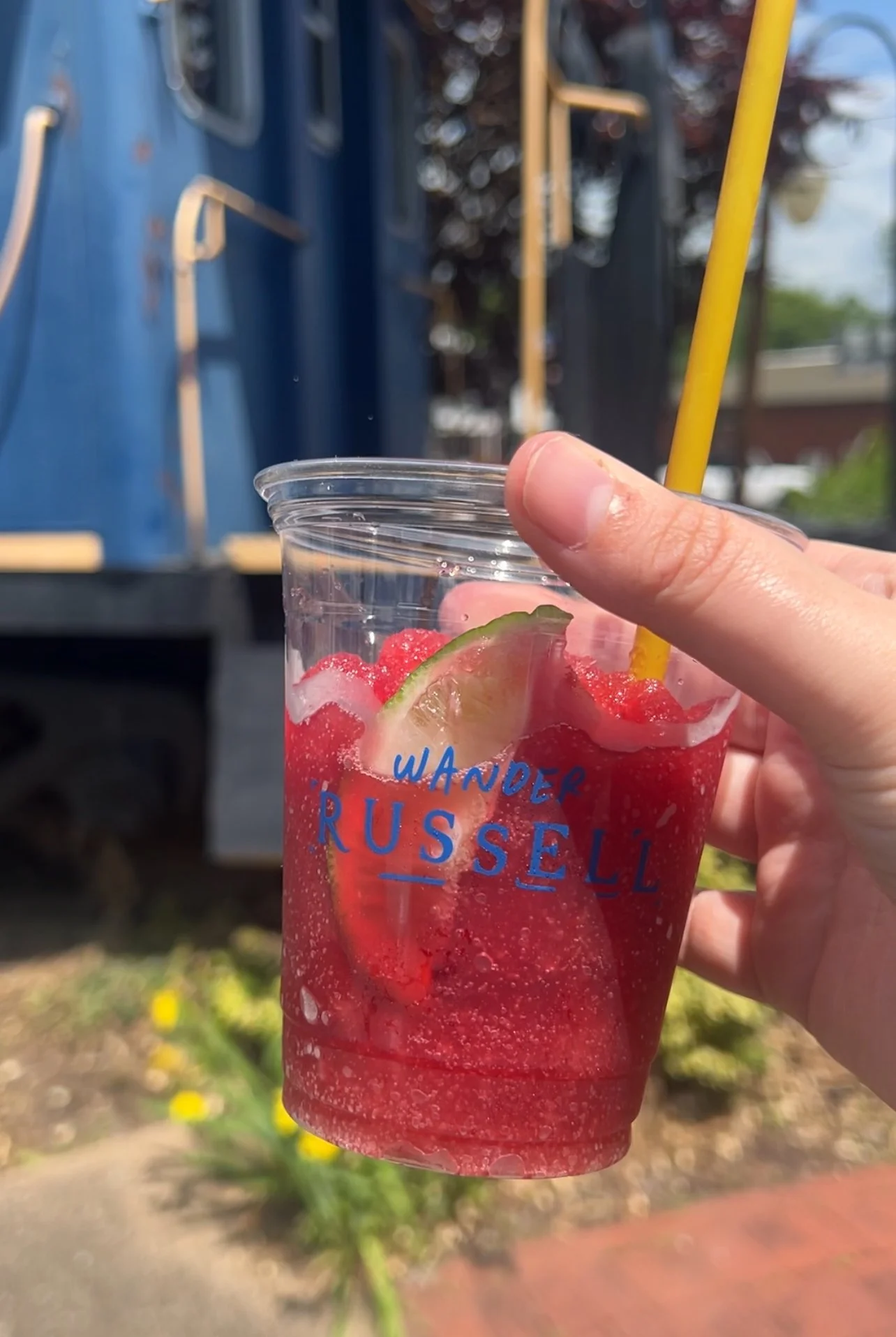 A clear plastic cup with blue text reading "WANDER RUSSELL," filled with a red soda, ice, lime slices, and a yellow straw, held outdoors.