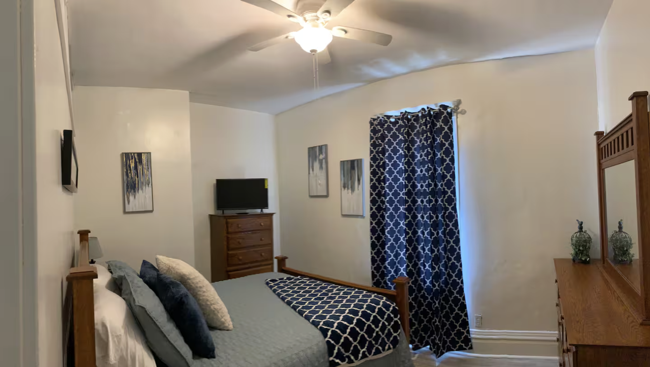 A bedroom with a wooden bed frame, gray and navy pillows, a gray bedspread, and a navy and white patterned blanket. There are two abstract paintings on the wall above the bed, a dresser with a TV on it, and a dresser with two decorative vases and a m