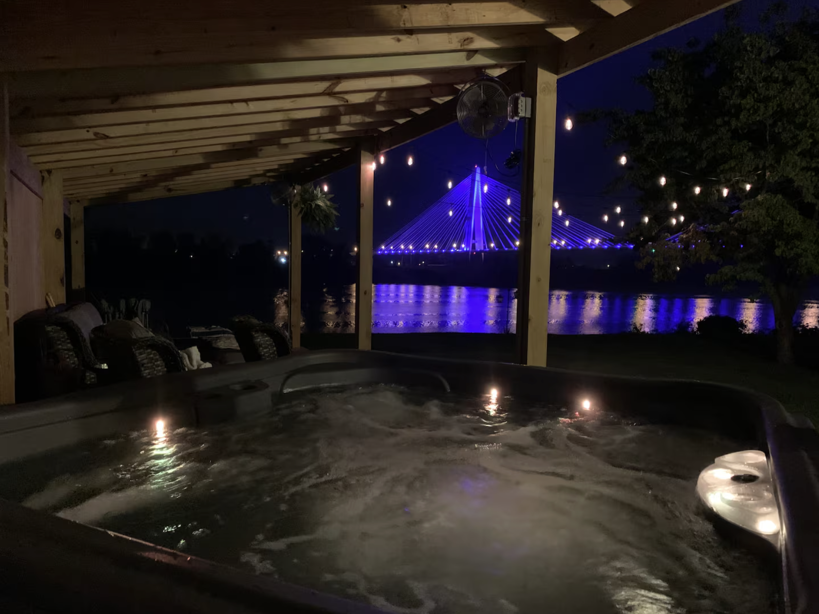 Night view from a hot tub on a covered porch overlooking a river with illuminated bridge in the background, string lights and a tree are visible.