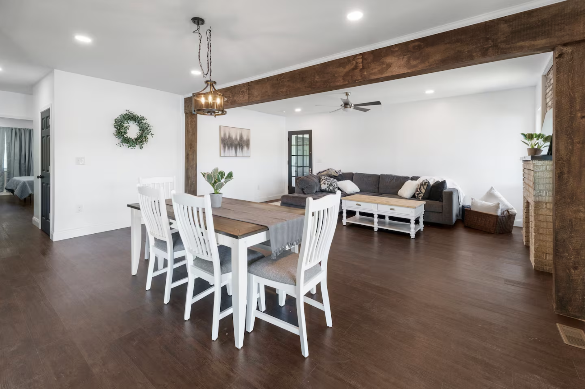 Open living and dining room with wooden floors, white walls, a dining table with four white chairs, a hanging light, a gray sectional sofa with pillows, wall art, a ceiling fan, and a brick fireplace with a plant on the mantel.