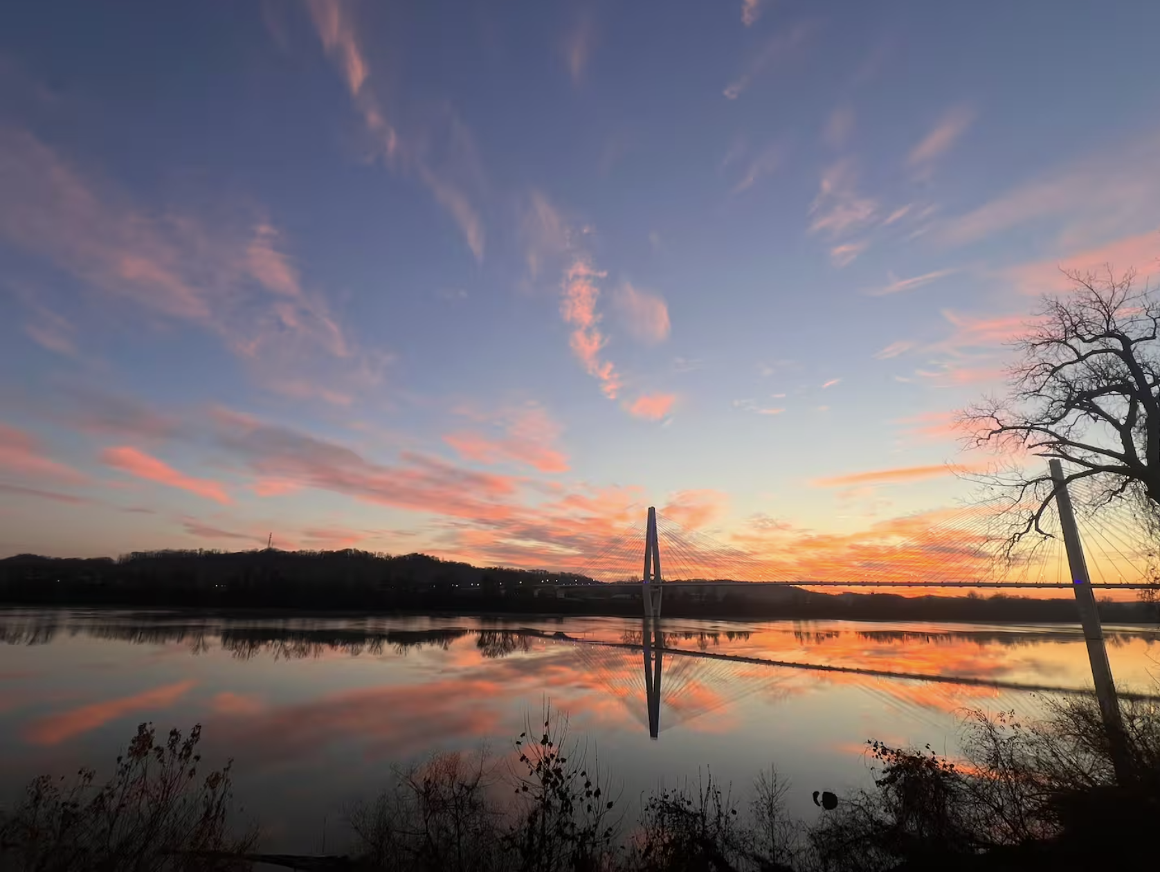 Sunset over a river with colorful sky, bridge, and silhouette of a leafless tree.