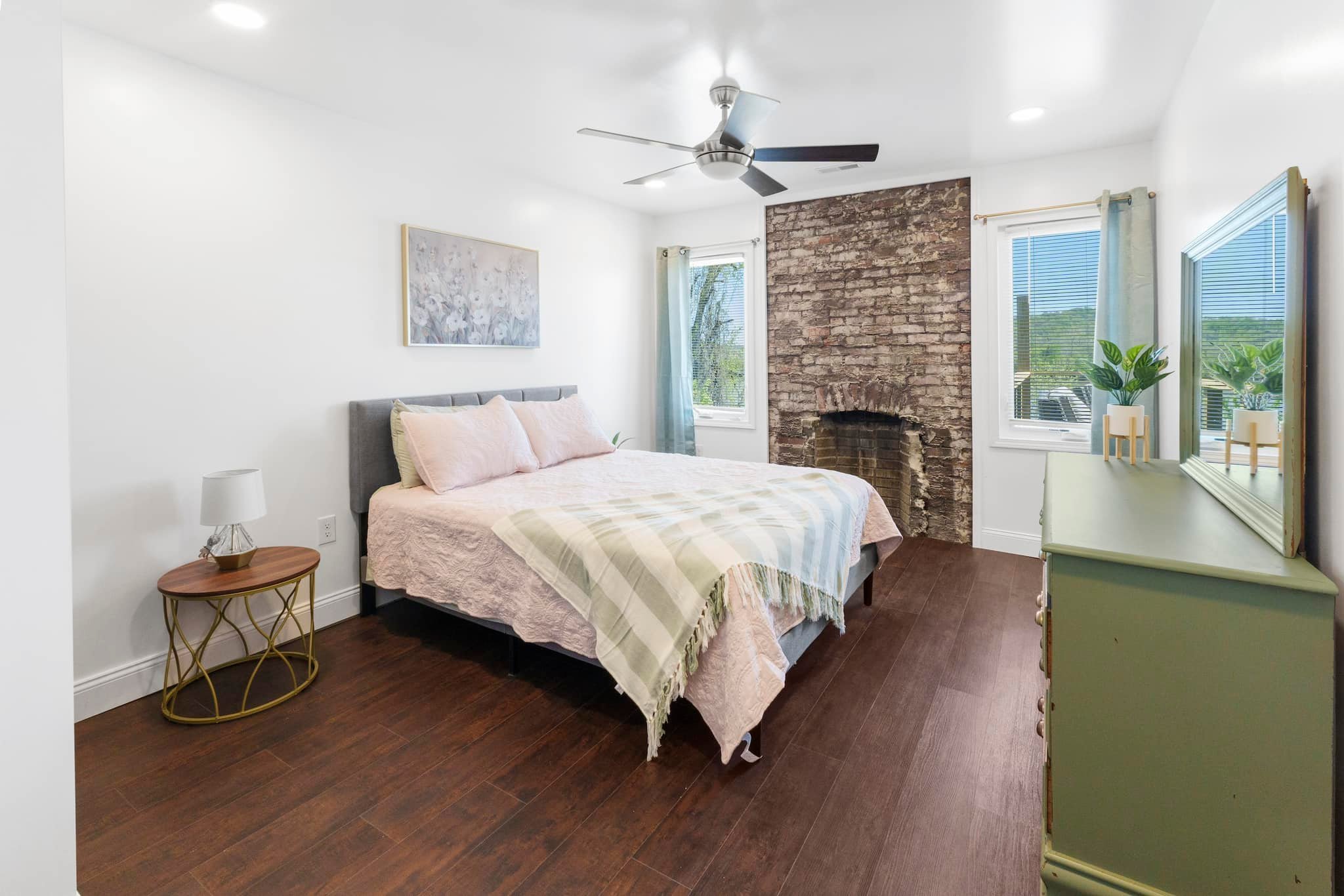 Bedroom with a bed, nightstand, exposed brick fireplace, dresser with mirror, two windows with curtains, ceiling fan, and hardwood floors.