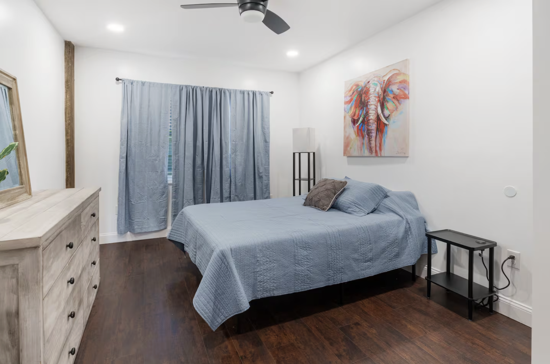 Bedroom with white walls, dark hardwood floor, blue bedspread, gray pillows, blue curtains, artwork of a colorful elephant on the wall, a wooden dresser with a mirror, a black bedside table, a tall lamp, and a ceiling fan.