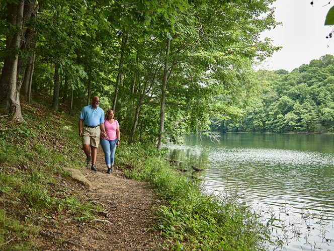 A man and woman walking along a forested trail beside a lake.