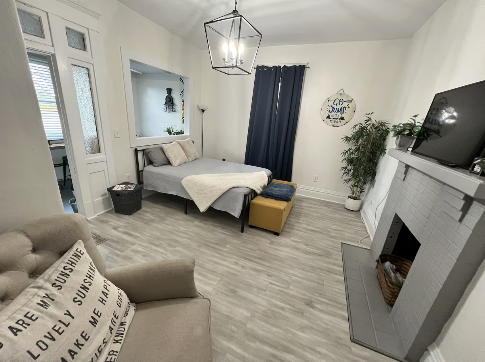 A cozy living room with a bed and a small dining area visible through an open door in the background. The room features a beige armchair with a decorative pillow, a white fireplace with a basket inside, a small ottoman near the bed covered with blank