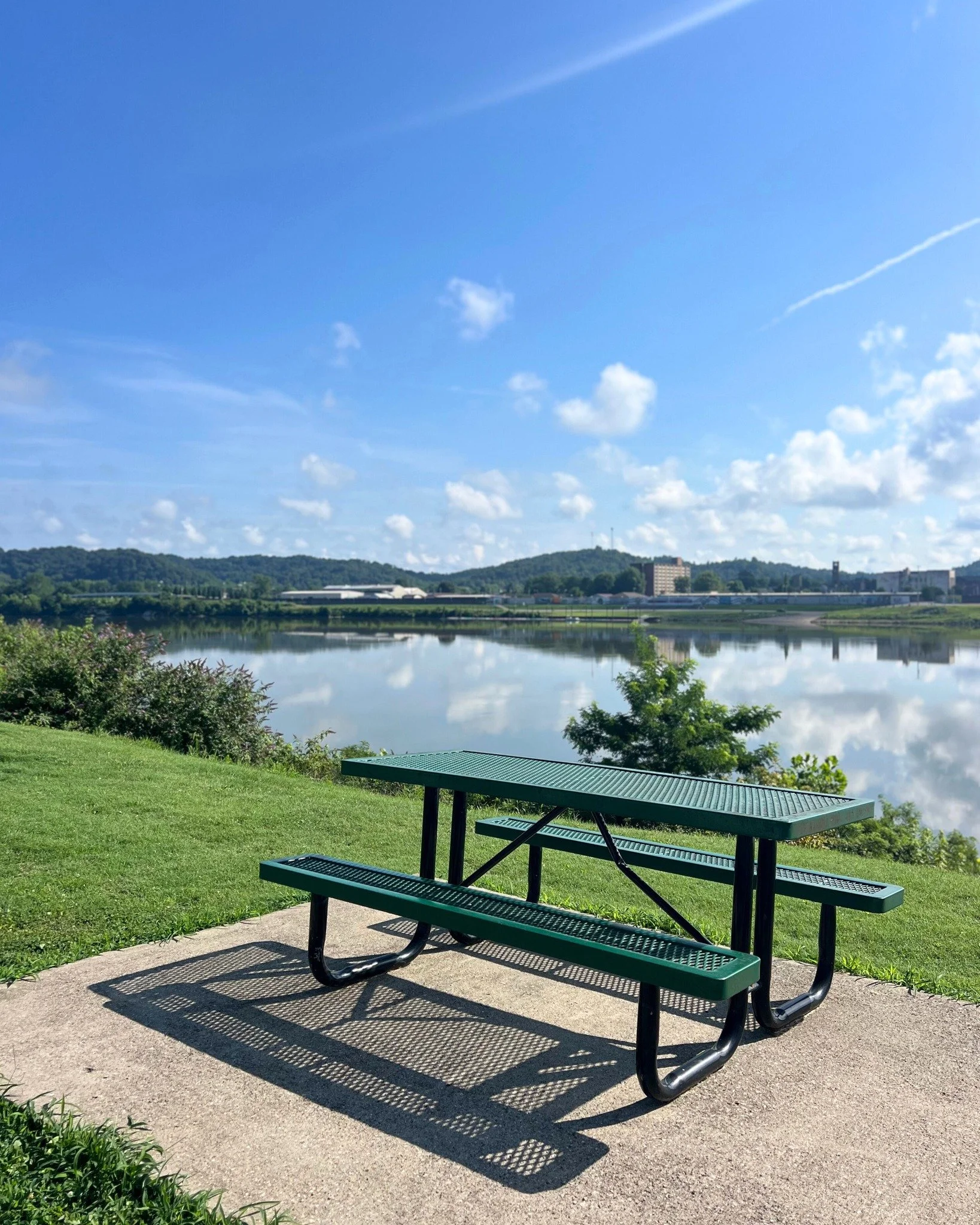 A green public picnic table on a concrete pad near a lake with a cityscape and hills in the background, under a partly cloudy blue sky.