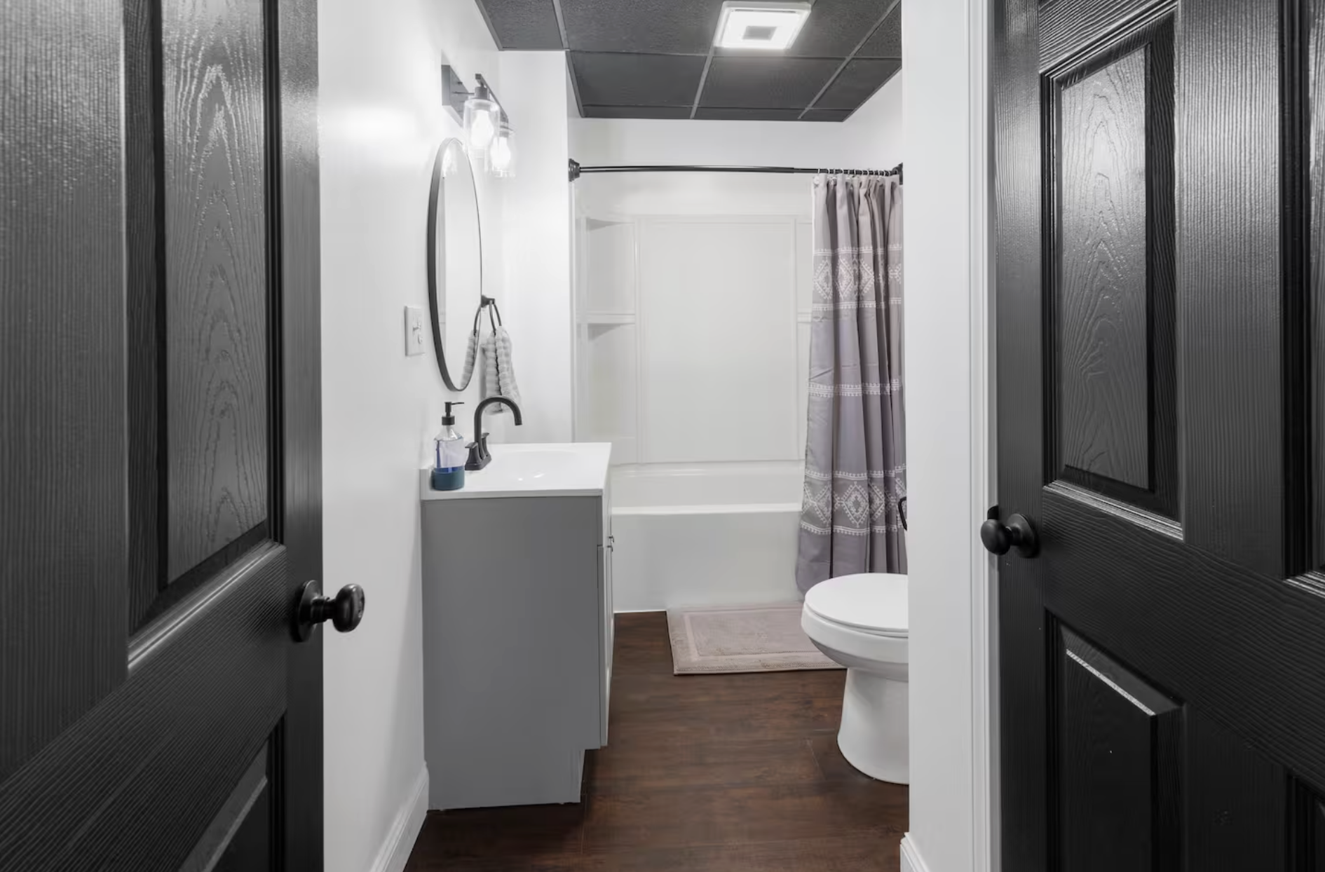View of a modern bathroom seen through open black doors, featuring a white vanity with a round mirror, a bathtub with a shower curtain, and a toilet, with wooden flooring and white walls.