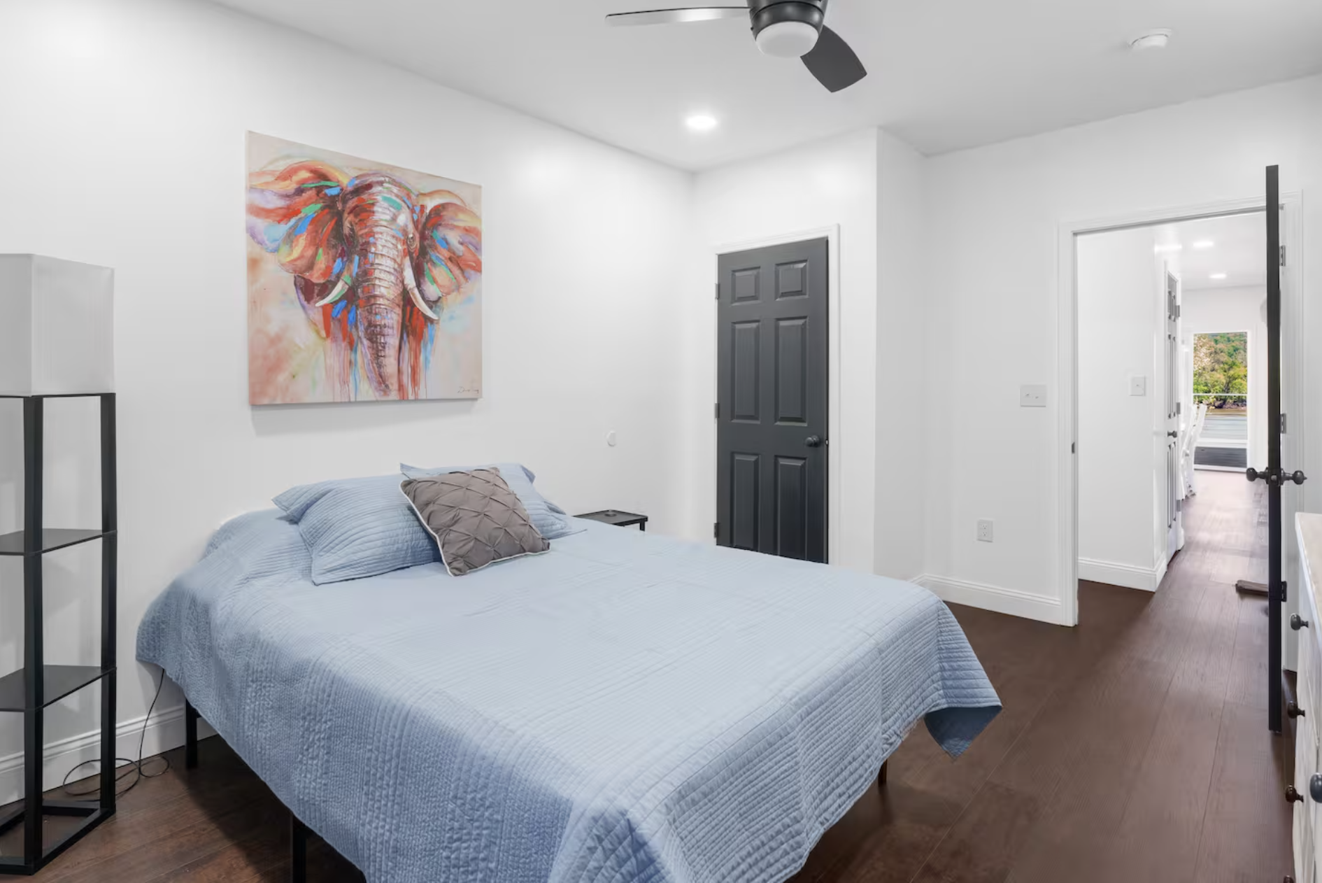Bedroom with a bed covered in a light blue quilt, a decorative pillow, a bedside table, a colorful watercolor elephant painting on white walls, dark door, ceiling fan, hardwood floors, doorway to a dining area with table and chairs, and an outside vi