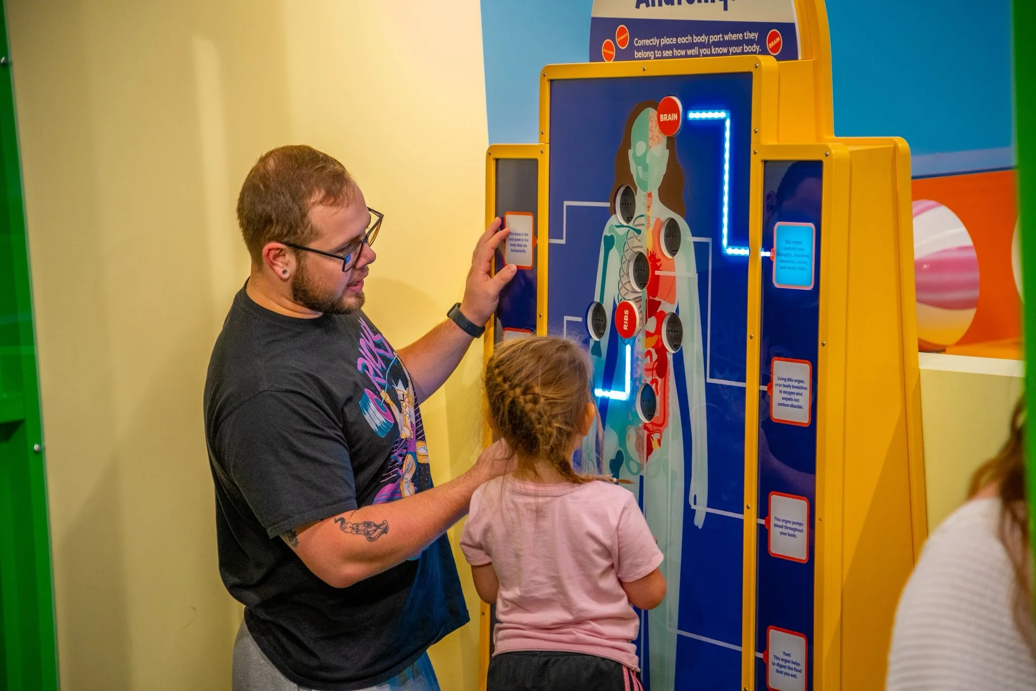 A man and a young girl participating in an interactive educational exhibit about the human body, with a diagram of a person and labeled body parts.