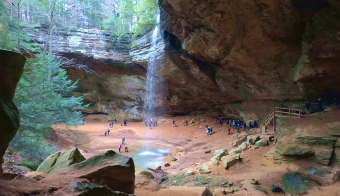 People exploring inside a Hocking Hills cave, a large sandstone alcove with a waterfall and a small pool, with a wooden observation platform on the right.