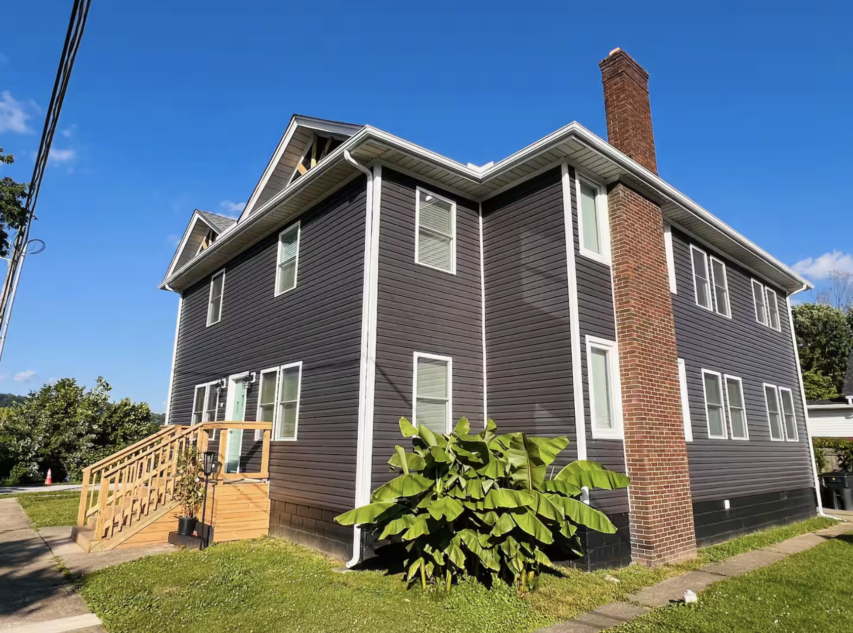 A two-story condo with dark siding, white trim, a brick chimney, and a small front porch with stairs, surrounded by green grass and plants under a clear blue sky.